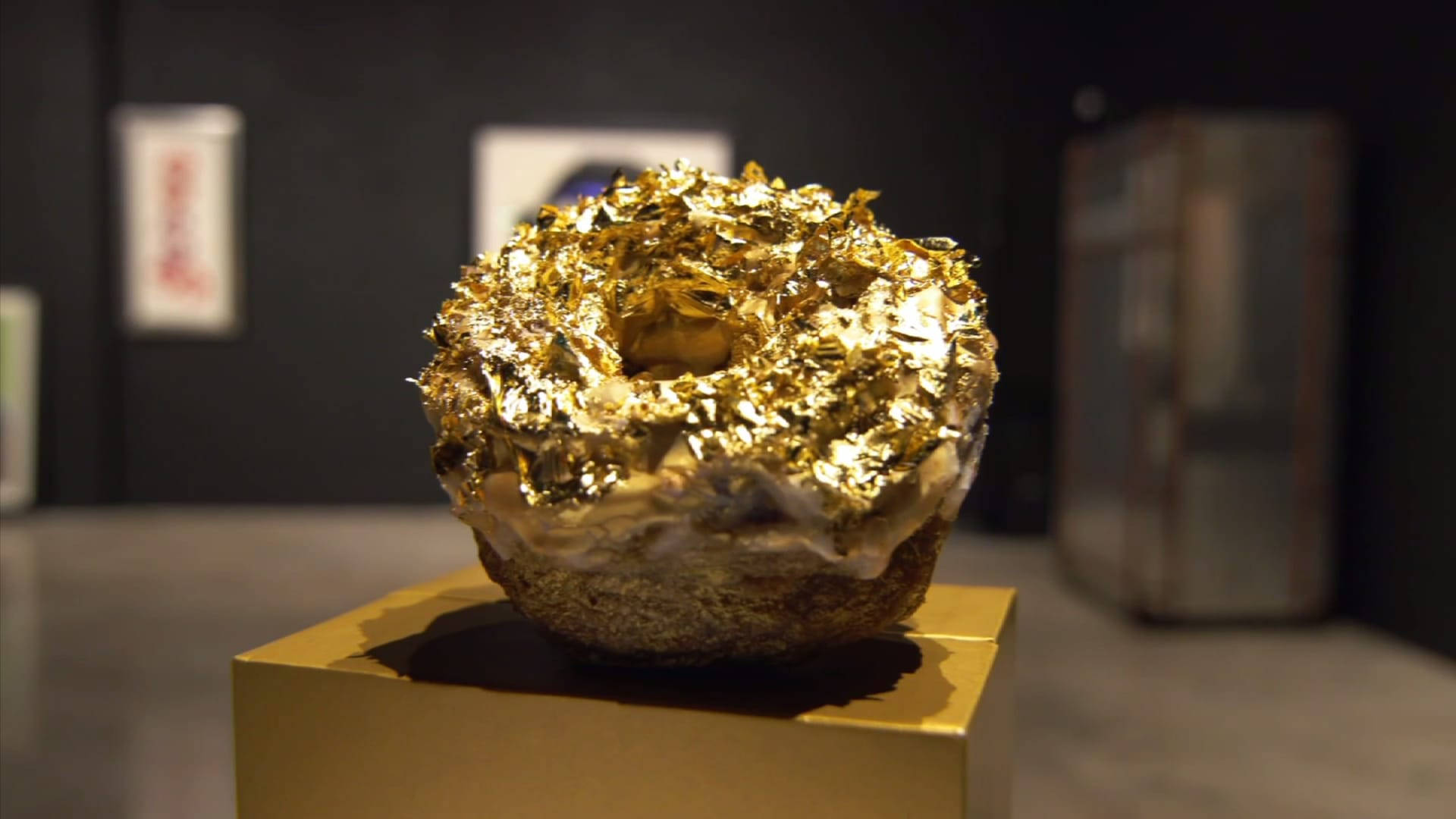 Foil 桌布 壁紙 — 一個覆蓋閃爍金箔的甜甜圈放在昏暗畫廊空間的金色底座上。