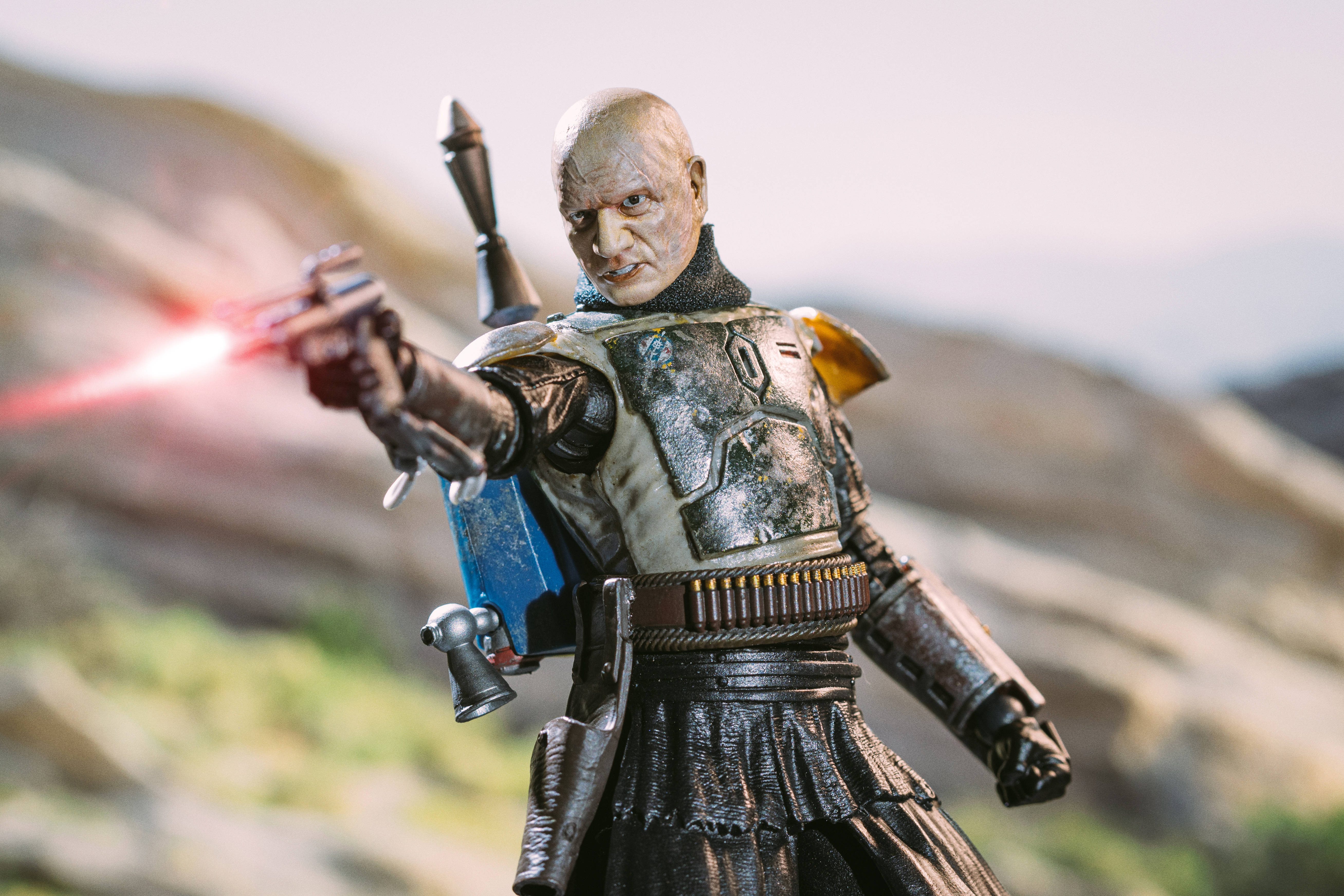 Boba 桌布 壁紙 — 一個詳細的動作人偶，描繪一位光頭戰士穿著風化盔甲，在模糊的岩石背景下發射紅色雷射手槍。