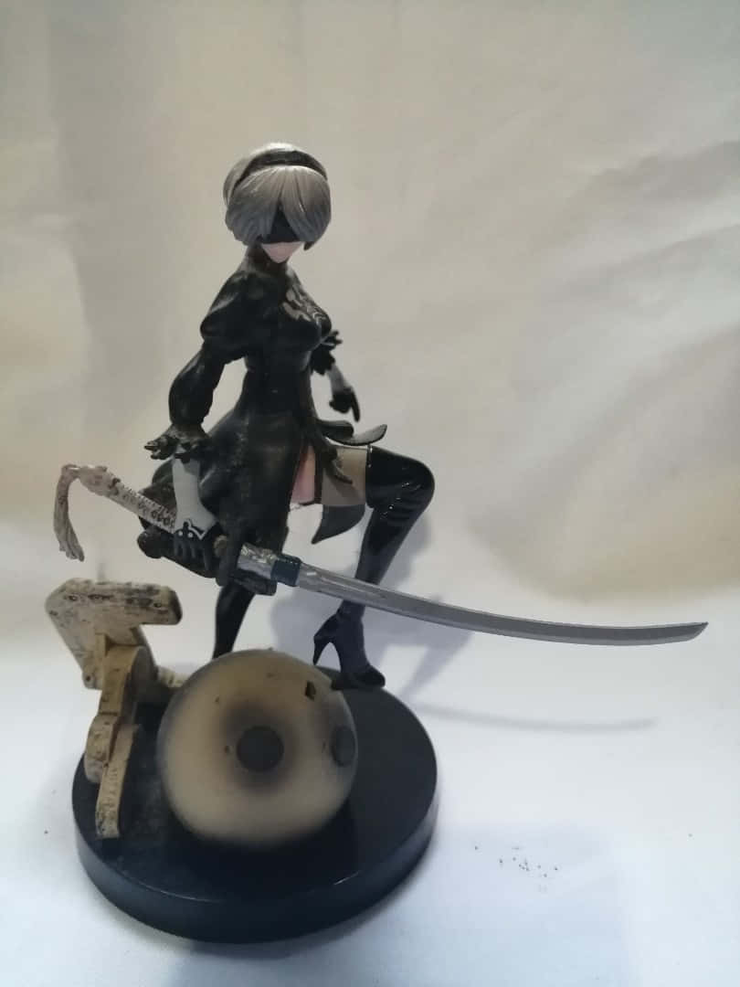 2B 桌布 壁紙 — 一個詳細的動漫人偶，短銀髮女戰士手持武士刀，站在帶有骷髏頭和木塊的黑色底座上。