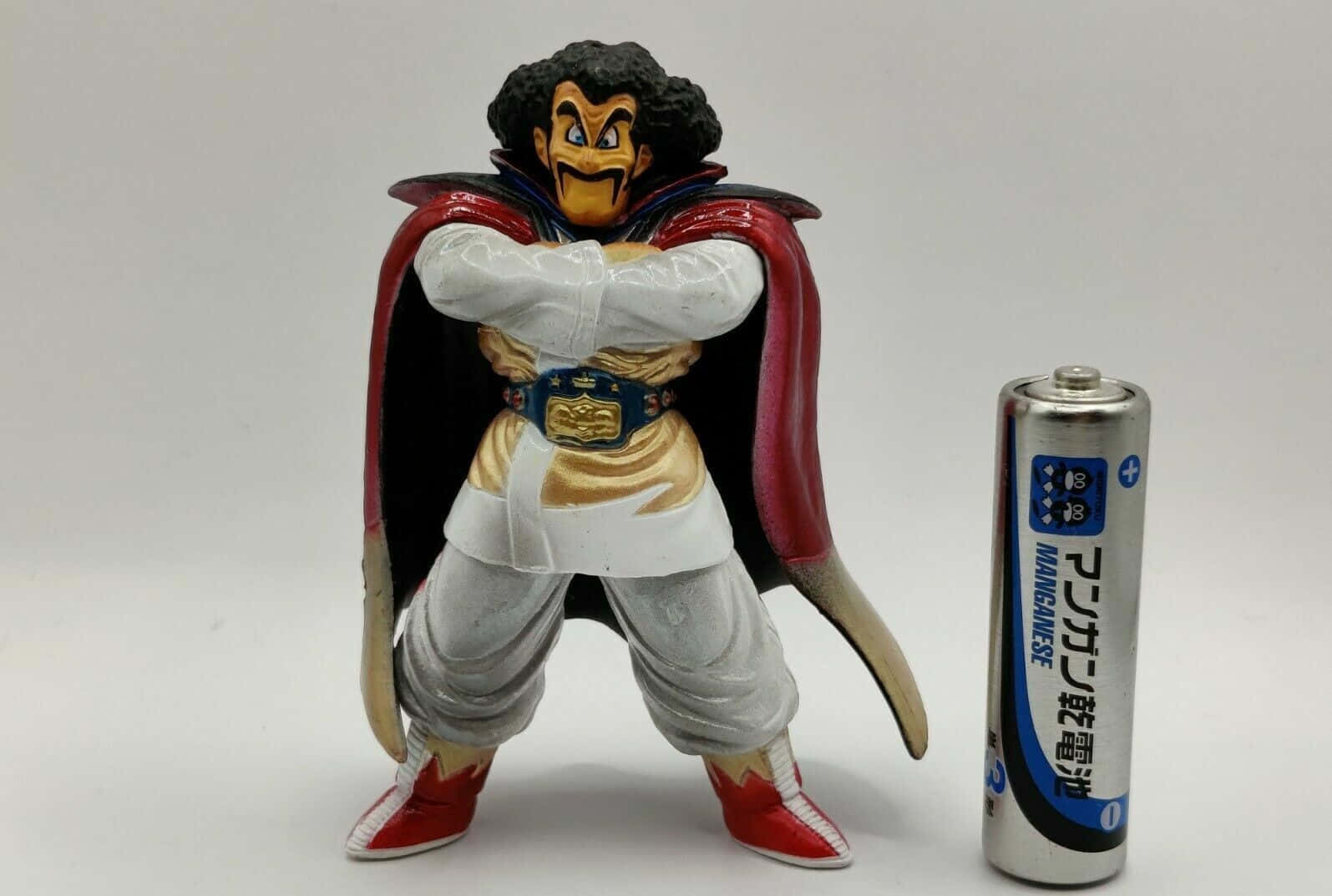 Hercule Satan 桌布 壁紙 — 一個詳細的動畫反派公仔，身穿紅色斗篷和金色腰帶，站在銀色電池旁邊，背景為純白色。