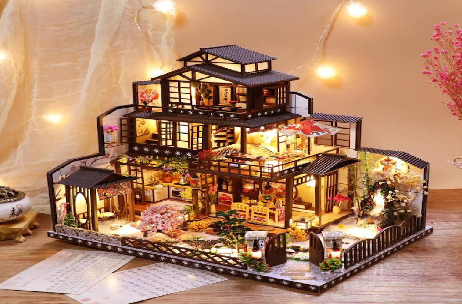 Dollhouse 桌布 壁紙 — 一個詳細的微型日式房屋，帶有溫暖的室內燈光和微型花園特色，展示在木質表面上。