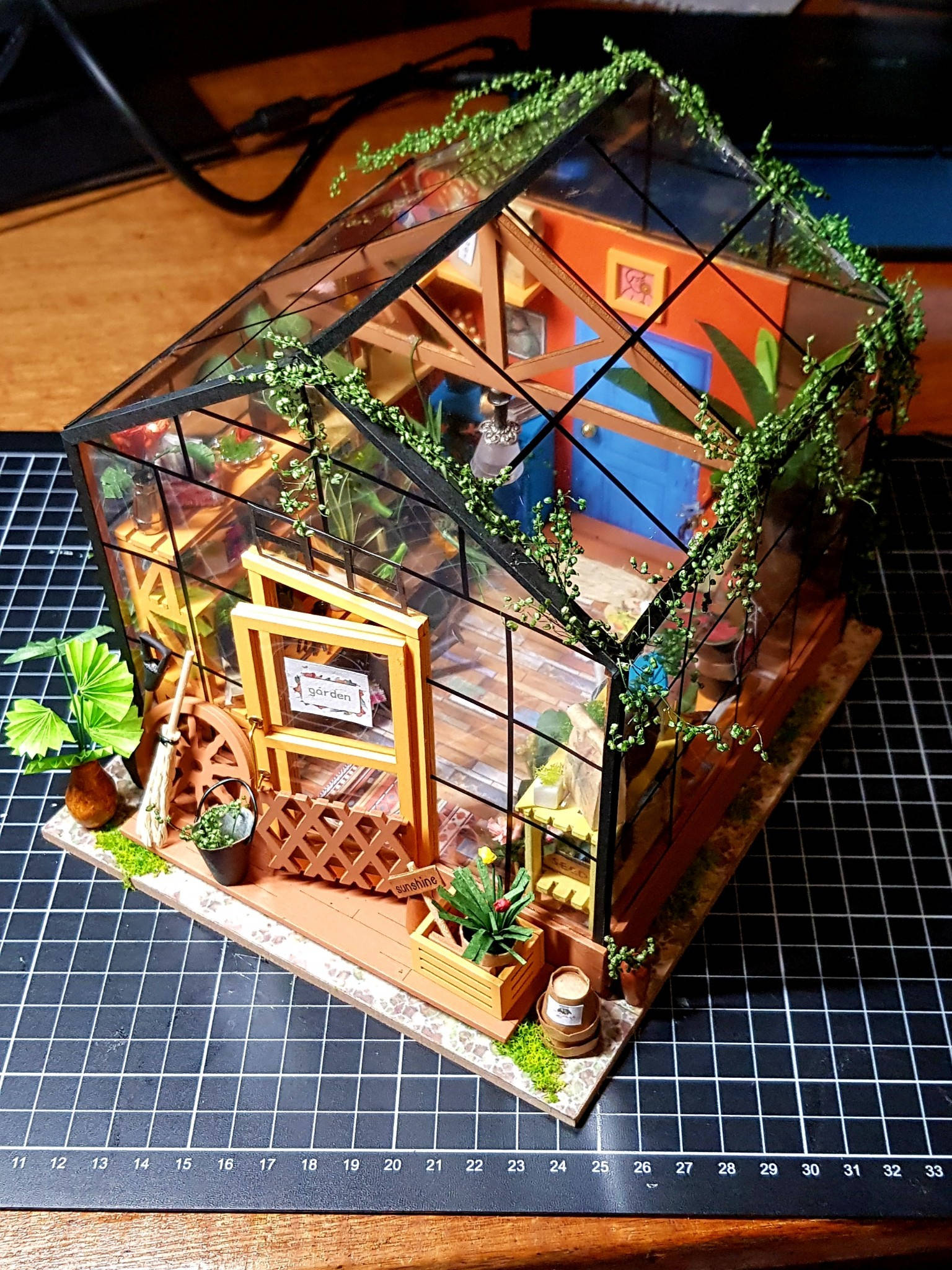 Dollhouse 桌布 壁紙 — 一個詳細的微型溫室模型，帶有玻璃面板、木框和內部茂盛的綠色植物，放置在網格切割墊上。