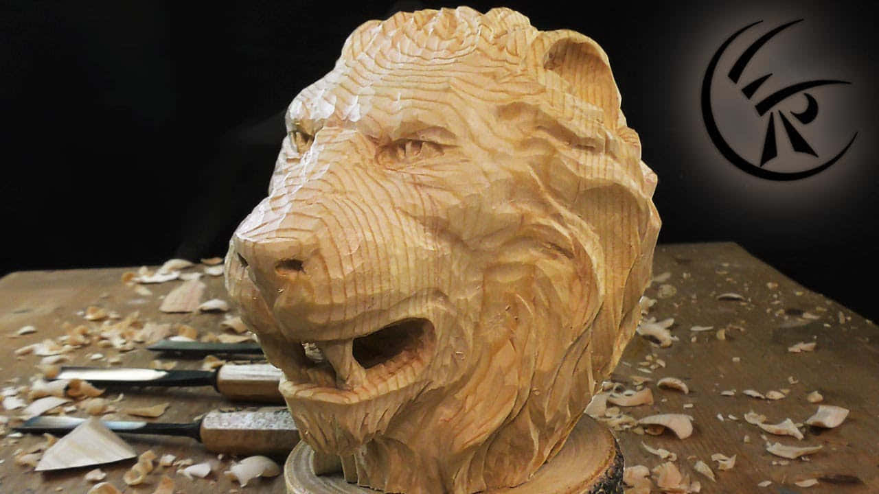 Lion Head 桌布 壁紙 — 一個詳細的木製獅子頭雕塑，帶有雕刻的鬃毛和張開的嘴巴，放在一個被木屑和雕刻工具包圍的工作台上。