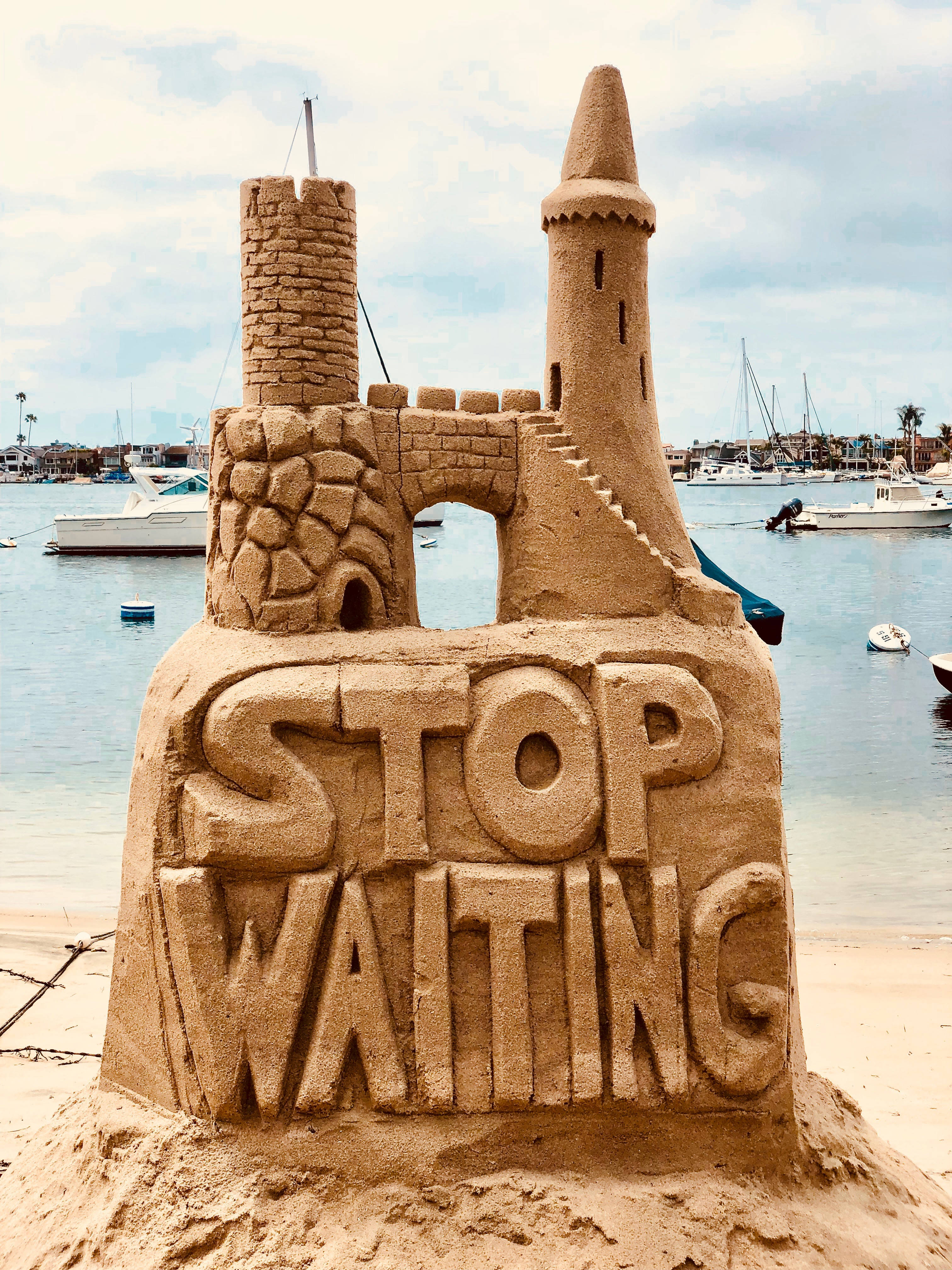 Quote 桌布 壁紙 — 一個詳細的沙堡，帶有城堡塔樓，底座上刻有顯眼的「STOP WAITING」字樣，背景是遊艇碼頭、船隻和寧靜的水面。