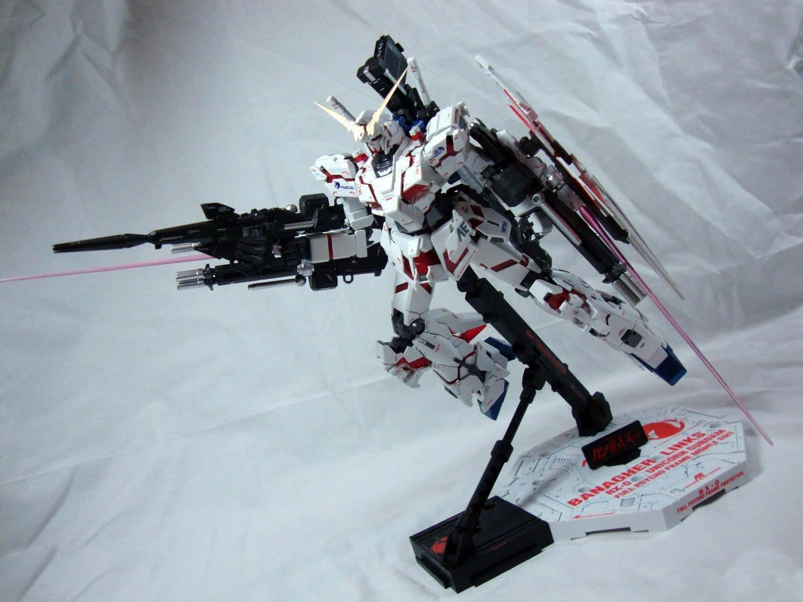 Gundam Unicorn 桌布 壁紙 — 一個詳細的白色、紅色和黑色鋼彈模型，在皺巴巴的白色背景前，動態地擺放在支架上。