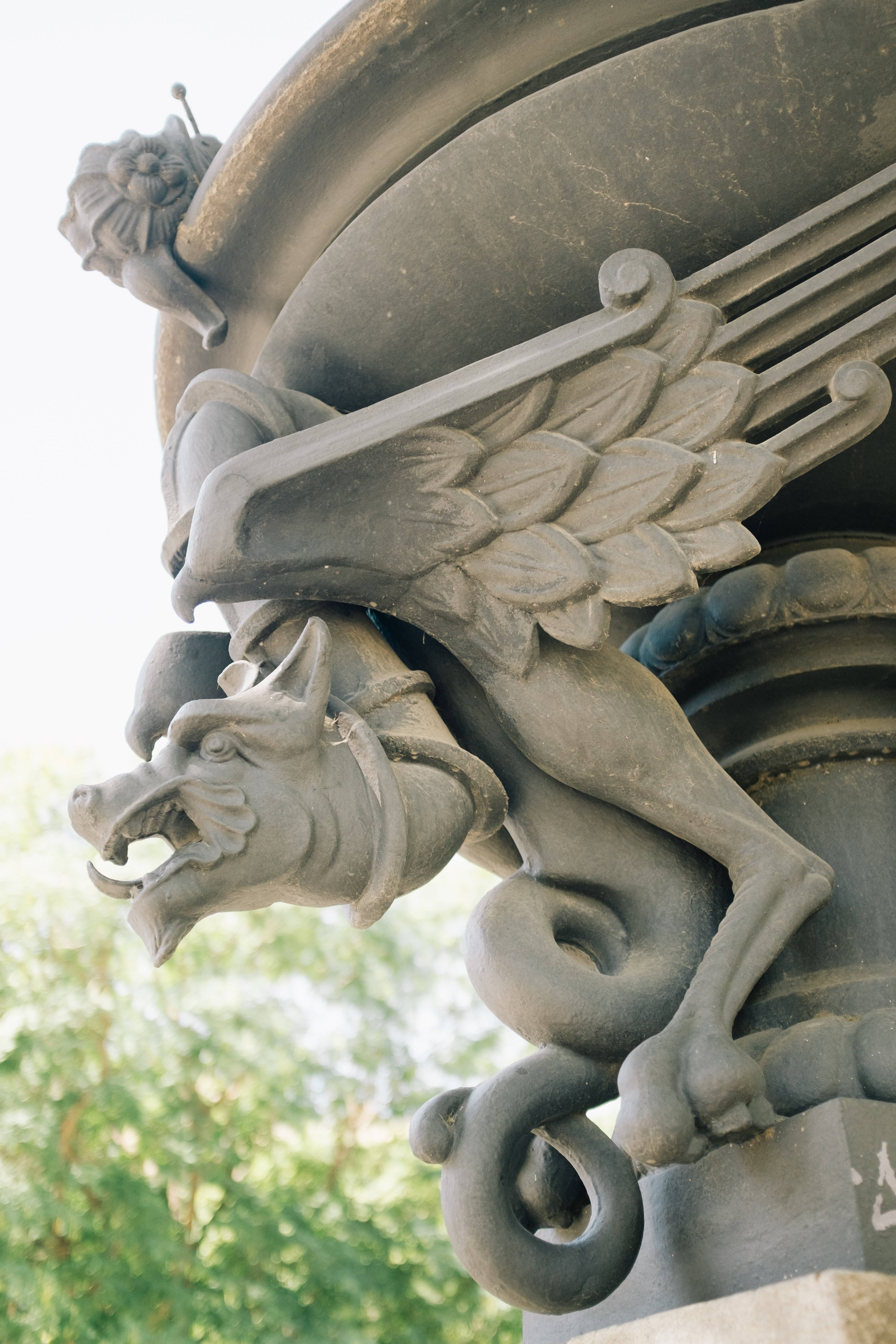 4K iPhone 6 plus 桌布 壁紙 — 一個詳細的石像鬼特寫，有翅膀和狗狀頭部，棲息在華麗的建築柱子上，背景是模糊的綠色樹葉。
