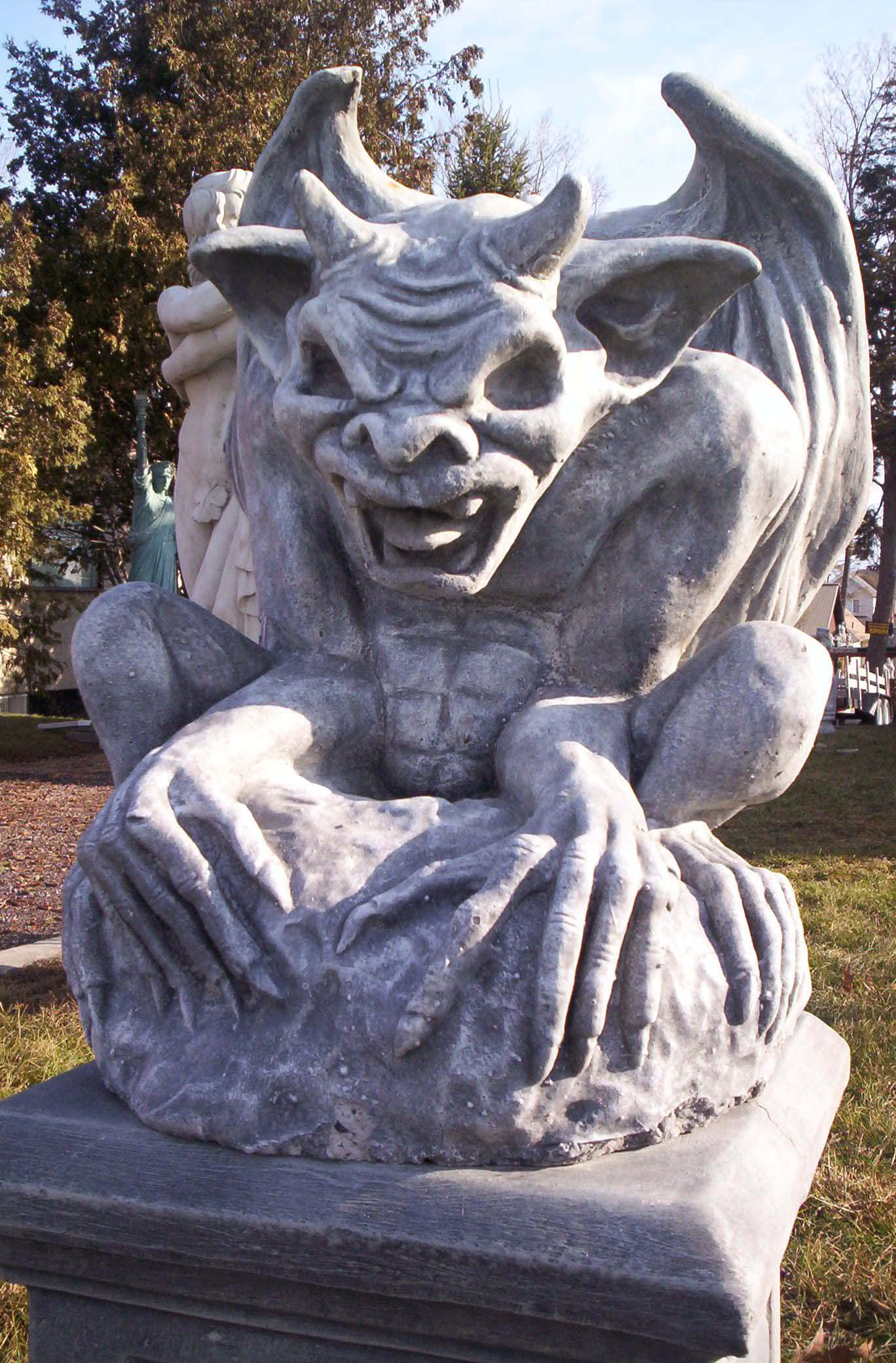 Gargoyle 桌布 壁紙 — 一個詳細的石像鬼雕像，有翅膀和角，威脅性地棲息在草地戶外環境的基座上。