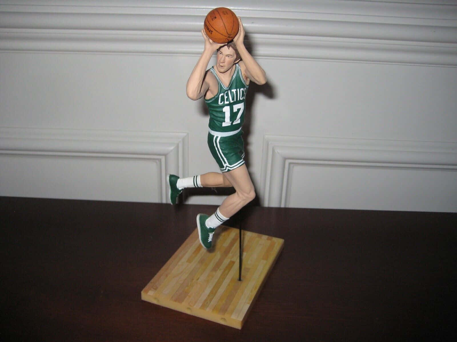 籃球 桌布 壁紙 — 一個詳細的籃球運動員人偶，身穿綠色塞爾提克隊17號球衣，準備投籃，站在木製底座上，背景是白色鑲板牆。