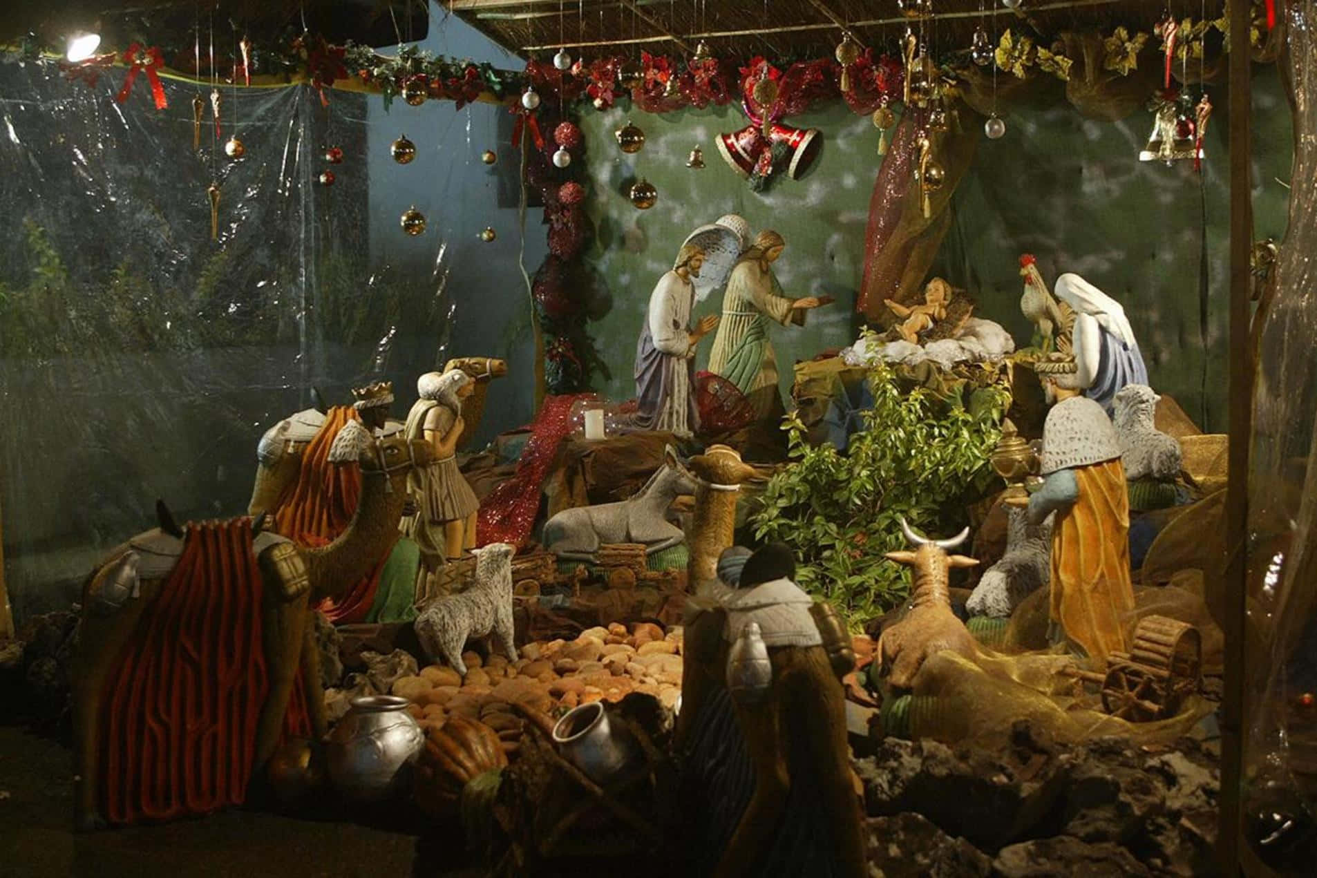 Religious Christmas 桌布 壁紙 — 一個詳細的耶穌誕生場景，包含瑪利亞、約瑟、嬰兒耶穌、牧羊人和動物的雕像，下方裝飾著節日紅色花環和懸掛飾物。