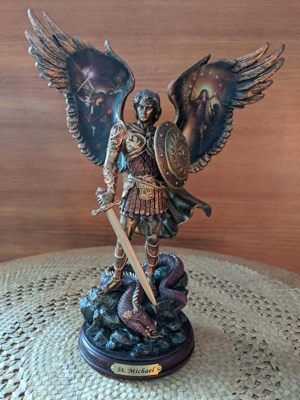 Saint Michael 桌布 壁紙 — 一個詳細的聖米迦勒天使長雕像，身穿盔甲，展開翅膀，勝利地站在一條龍之上，置於編織墊上。