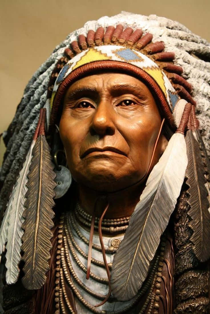 American Indian 桌布 壁紙 — 一個詳細的雕塑，描繪一位原住民長老，戴著羽毛頭飾和珠串項鍊，表情莊嚴。