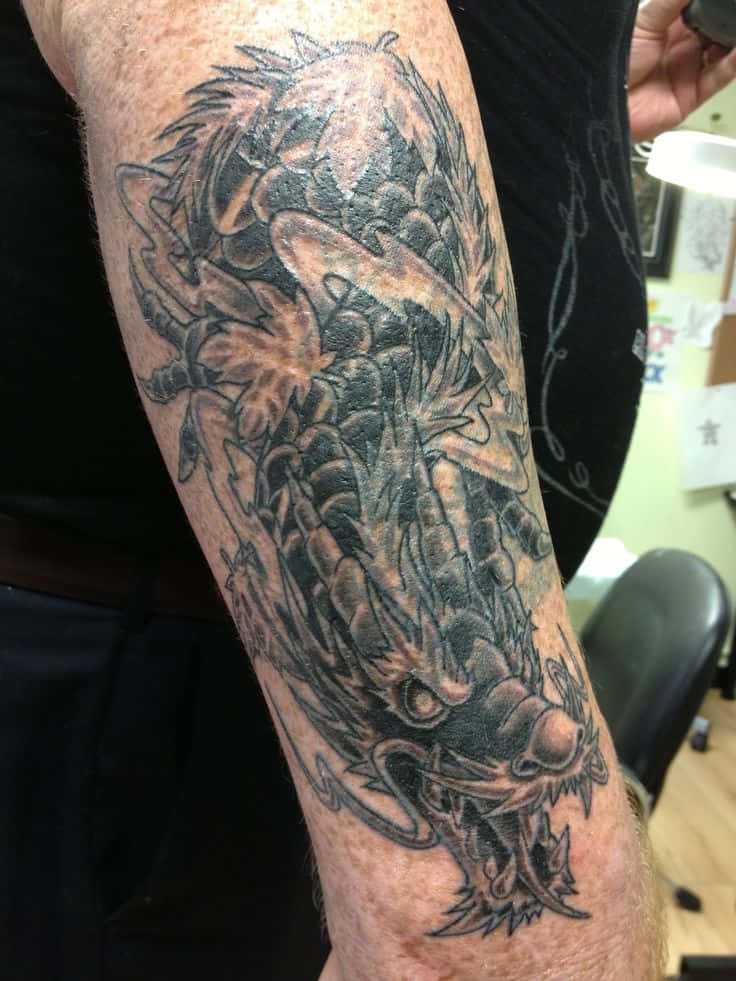 Tattoo 桌布 壁紙 — 一個詳細的黑灰色龍紋身，帶有鋒利的鱗片和兇猛的表情，戲劇性地纏繞在一個人的前臂上。
