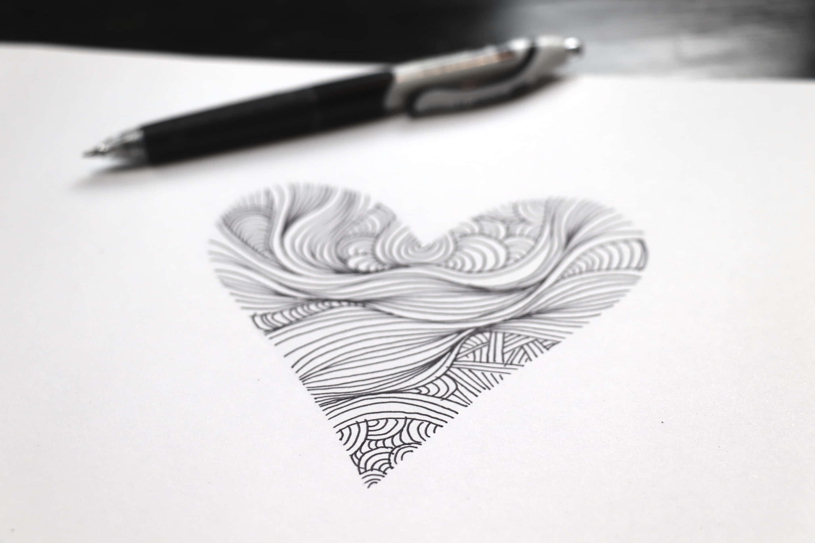 Heart Doodle 桌布 壁紙 — 一個詳細的黑白心形塗鴉，帶有複雜的漩渦圖案，放在白紙上，旁邊有一支黑色筆。