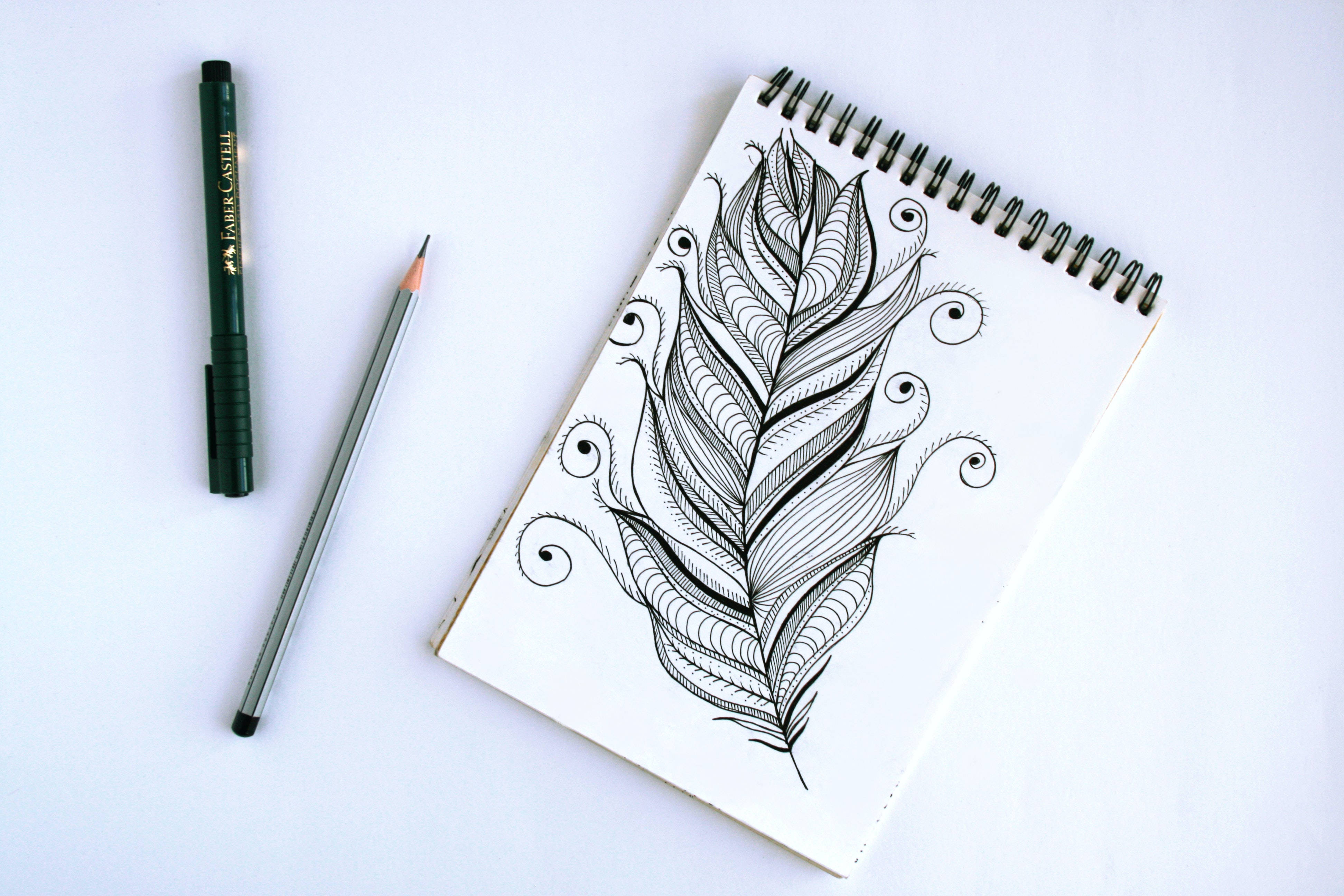 pencil 桌布 壁紙 — 一個詳細的黑色墨水羽毛塗鴉填滿了一本螺旋筆記本，旁邊有一支綠色鋼筆和鉛筆，放在乾淨的白色表面上。