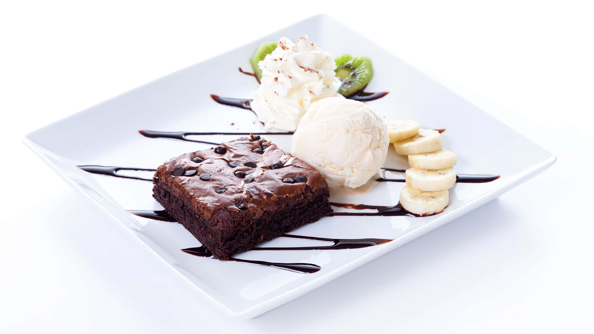 Desserts 桌布 壁紙 — 一個豐富的巧克力布朗尼，上面放有巧克力豆，旁邊有香草冰淇淋、奇異果片和香蕉片，淋上巧克力醬，放在白色盤子上。