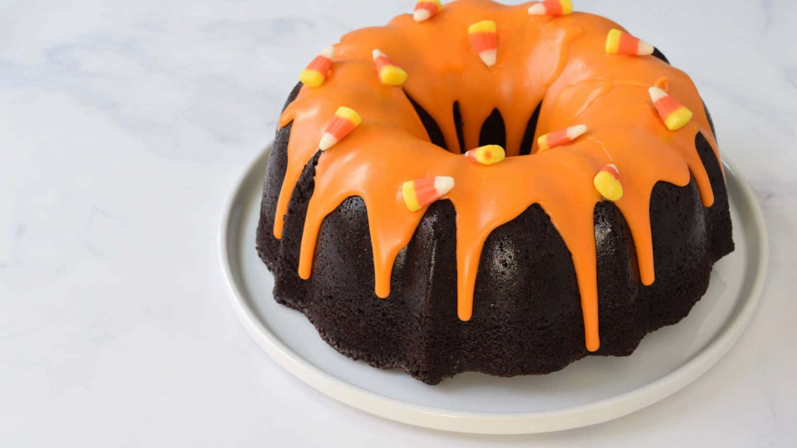 Halloween Cake 桌布 壁紙 — 一個豐富的巧克力環形蛋糕，淋上充滿活力的橙色糖霜，並在白色盤子上放上糖果玉米。