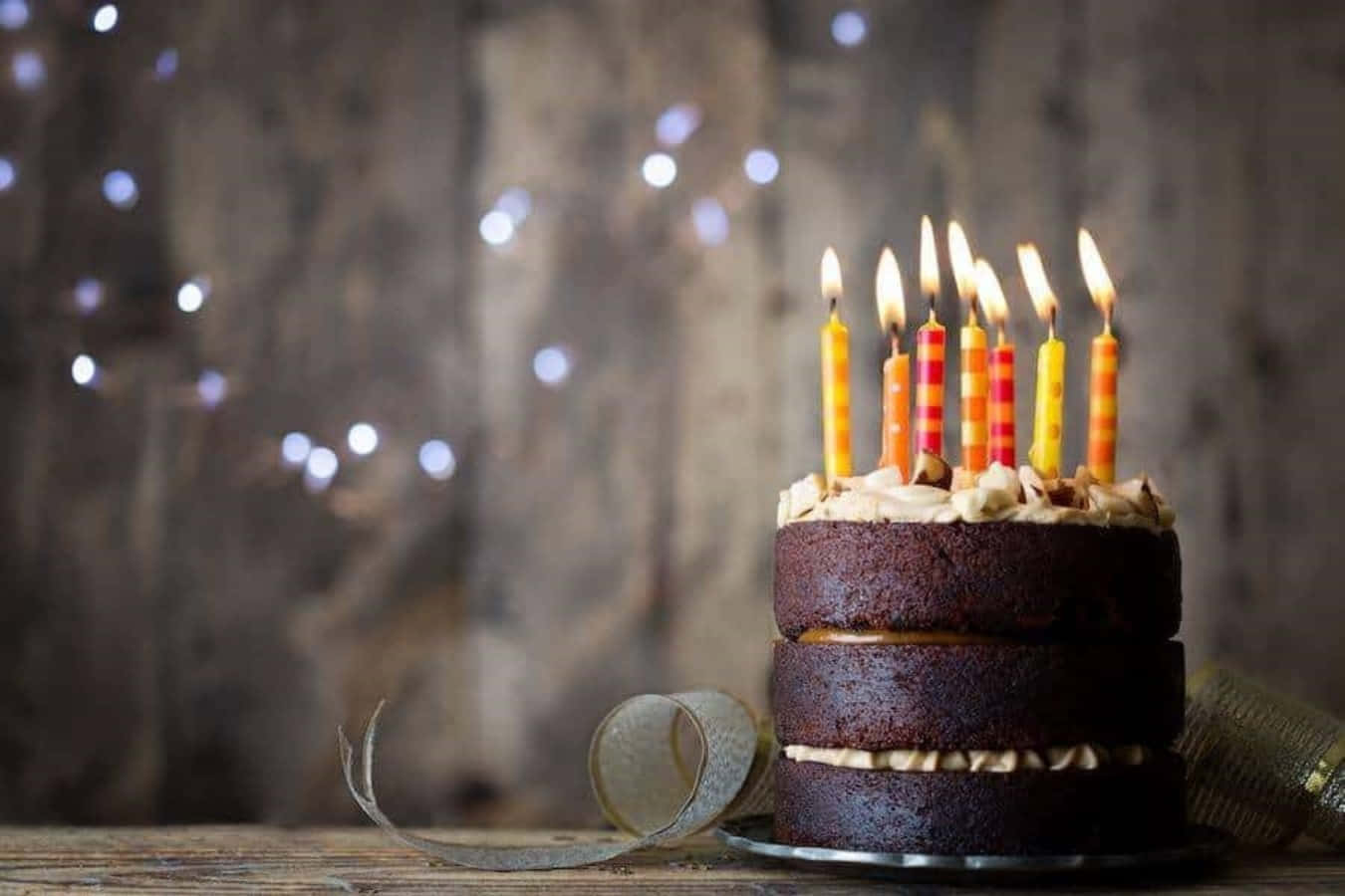 Birthday Celebration 桌布 壁紙 — 一個豐富的巧克力生日蛋糕，上面插著點燃的橙色和紅色蠟燭，放在一張質樸的木桌上，背景是柔和模糊的閃爍仙女燈。