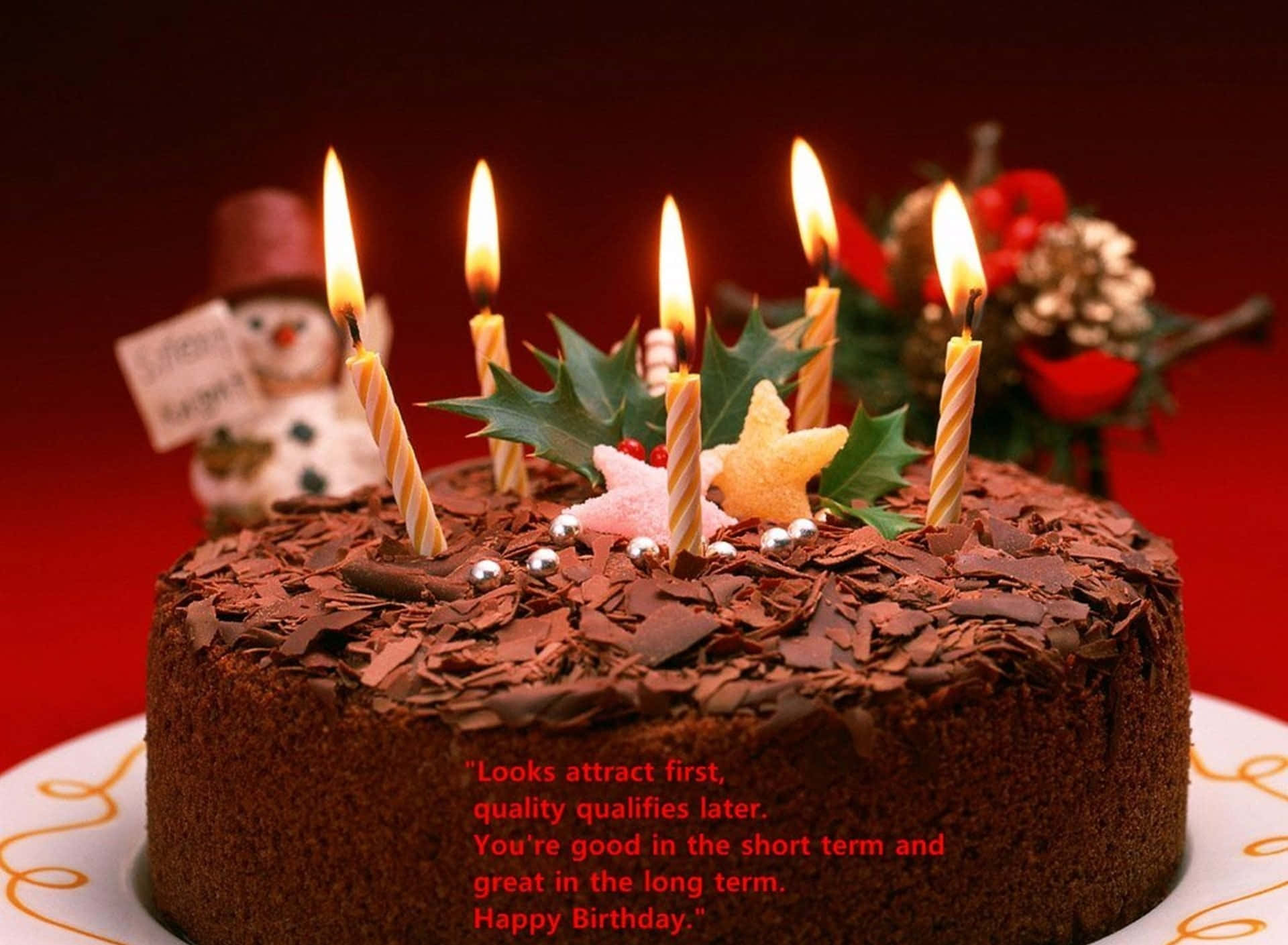 Birthday Wishes 桌布 壁紙 — 一個豐富的巧克力生日蛋糕，上面有燃燒的蠟燭、冬青果和雪人公仔，背景是深紅色。