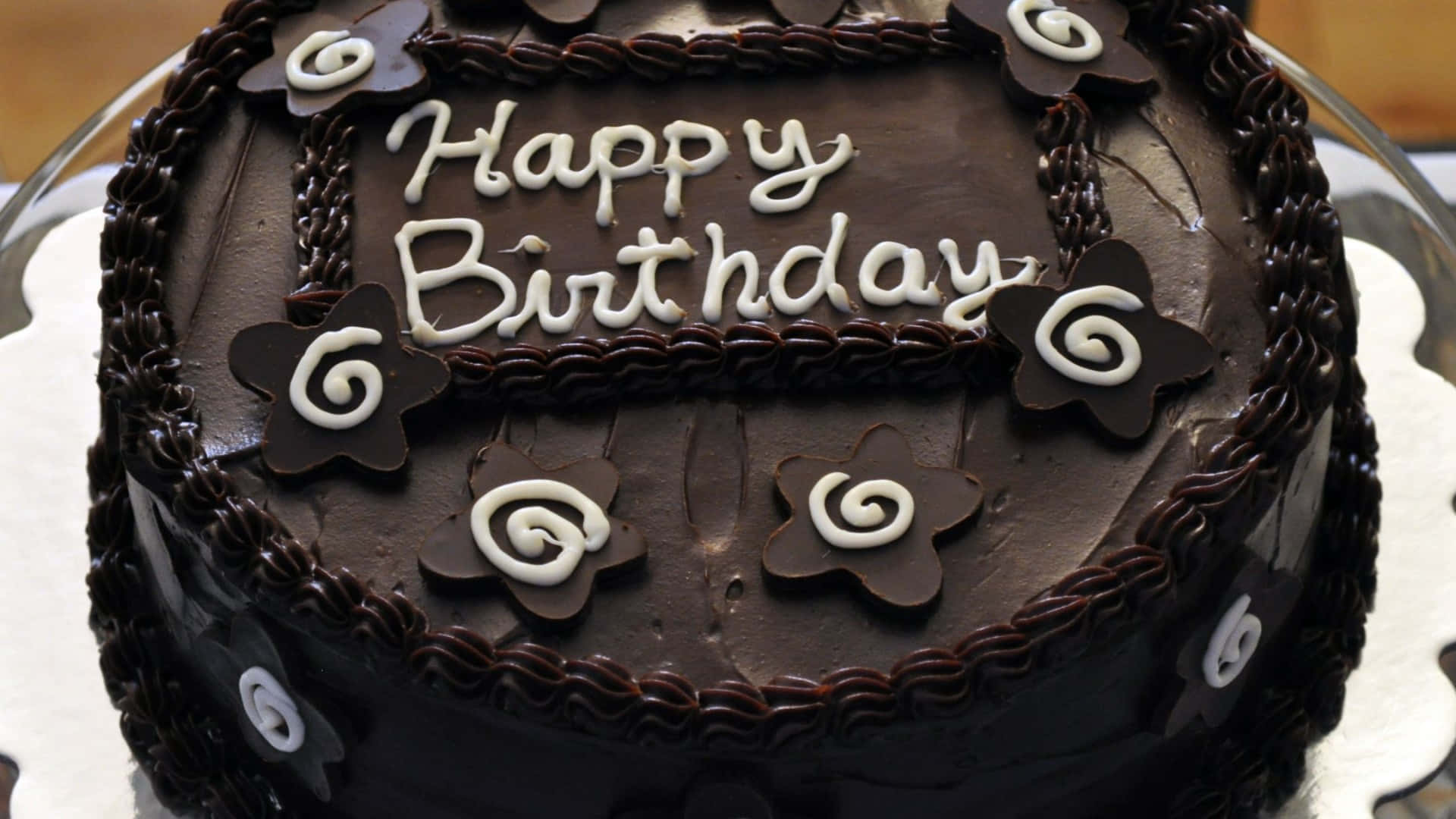 Birthday Cakes 桌布 壁紙 — 一個豐富的巧克力生日蛋糕，飾有白色糖霜文字和星形漩渦裝飾。