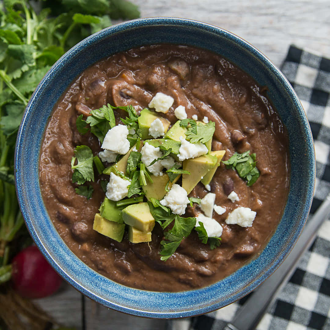 Black Bean Soup 桌布 壁紙 — 一個豐富的棕色豆燉菜，上面放有酪梨塊、碎起司和新鮮香菜，盛在藍色陶瓷碗中。