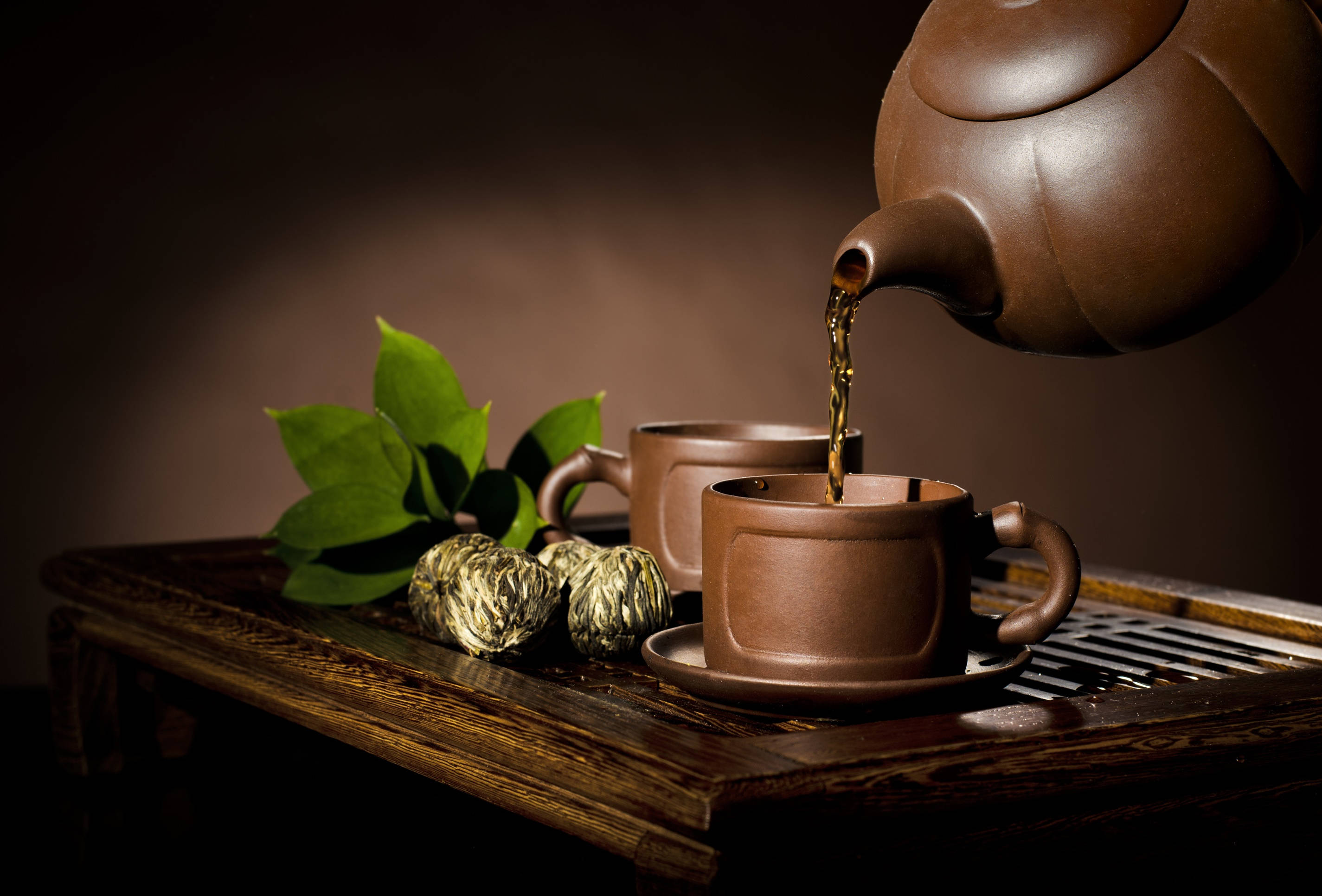 tea 桌布 壁紙 — 一個豐富的棕色陶壺將深色茶倒入木托盤上配套的杯子中，旁邊有綠葉和壓縮茶球。