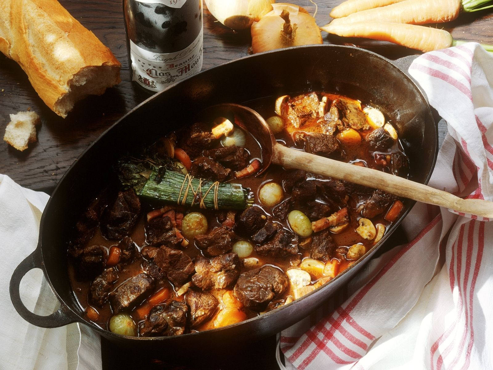 meat 桌布 壁紙 — 一個豐富的燉牛肉在鑄鐵鍋中慢煮，周圍是新鮮的胡蘿蔔、洋蔥和脆皮麵包，放在質樸的木桌上。
