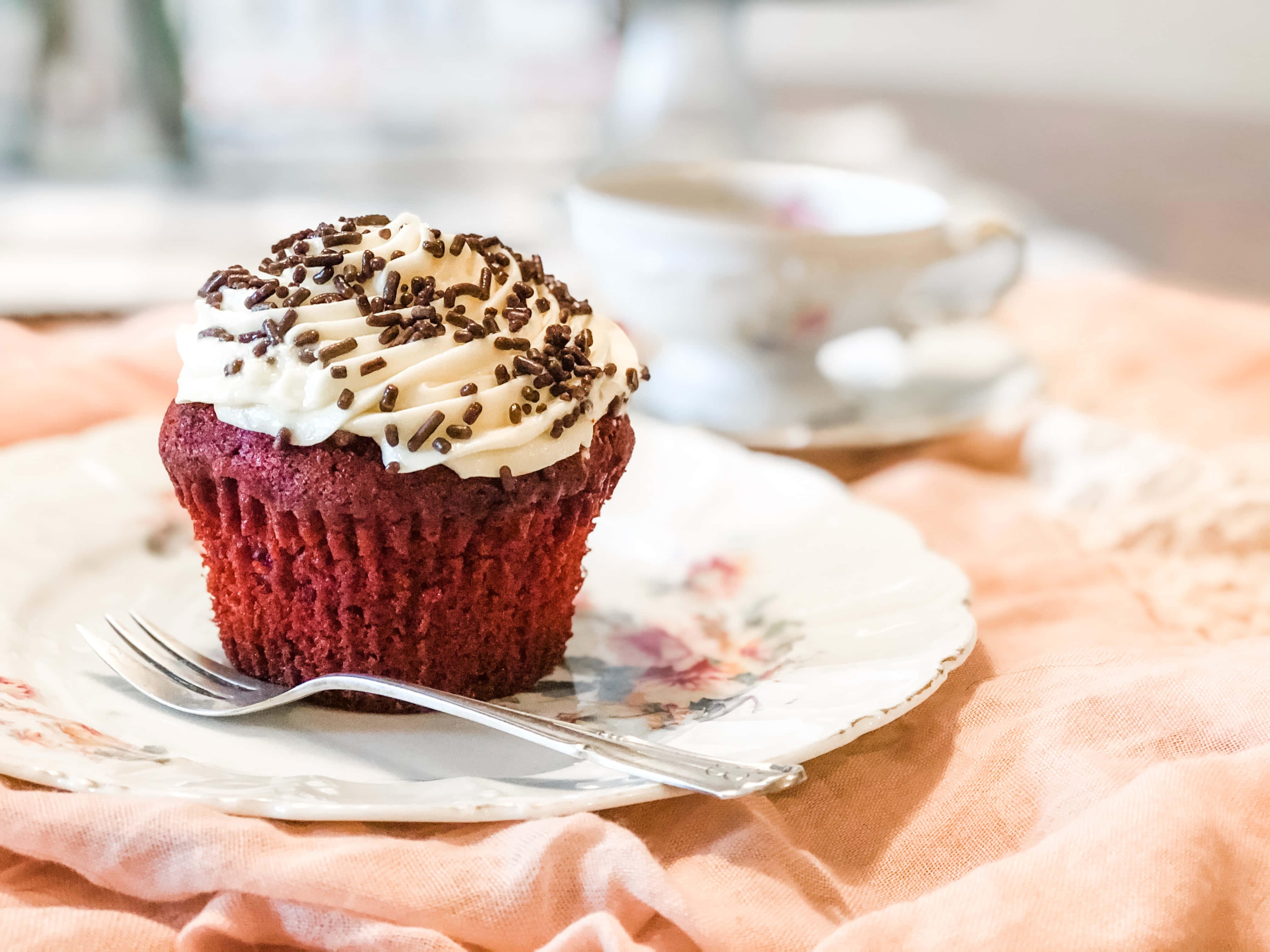 Cupcake 桌布 壁紙 — 一個豐富的紅絲絨杯子蛋糕，上面有奶油白色糖霜和巧克力彩針，放在一個花卉圖案的盤子上，旁邊有一個茶杯。
