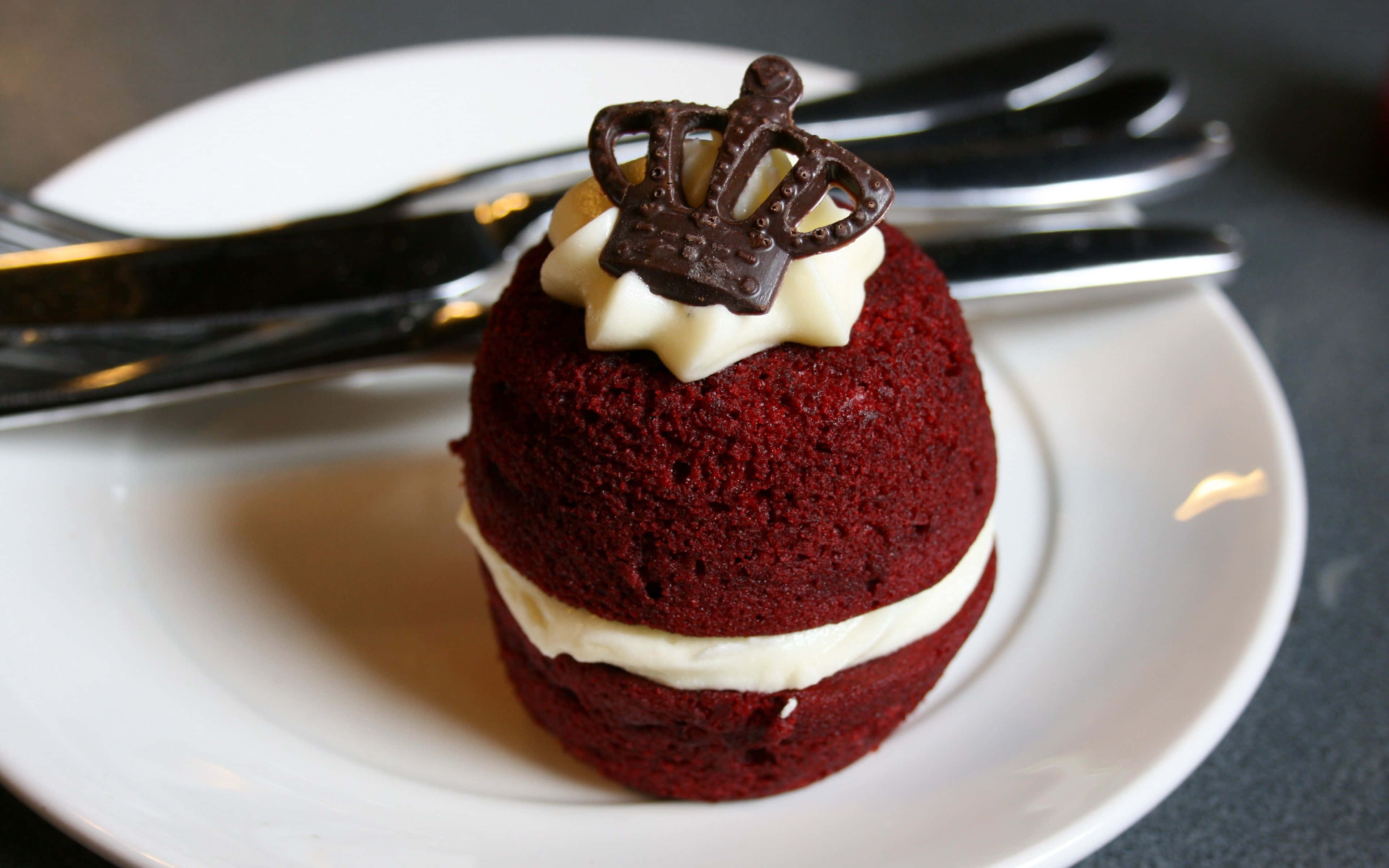 Red Velvet Cake 桌布 壁紙 — 一個豐富的紅絲絨杯子蛋糕，內有白色奶油餡料和巧克力皇冠，優雅地放在一個白色盤子上，旁邊有銀叉。