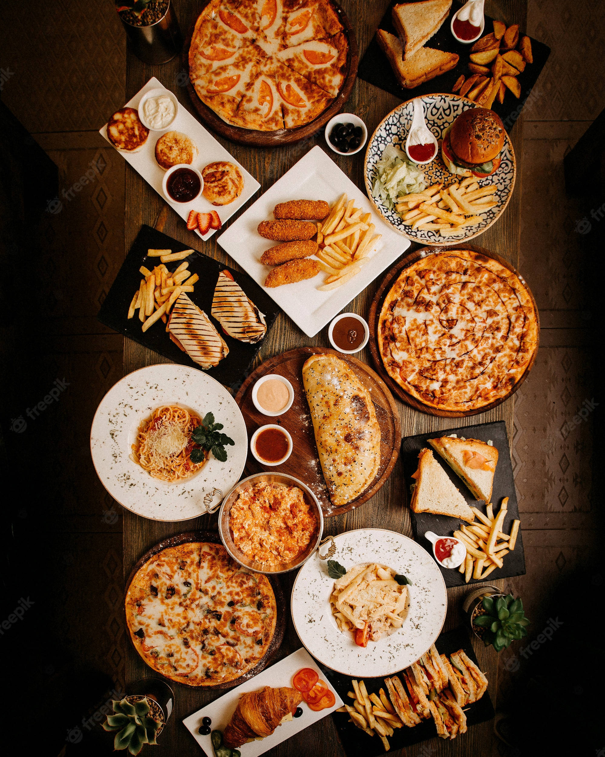 Pizza 桌布 壁紙 — 一個豐盛的俯視圖，展示各種快餐菜餚，包括披薩、漢堡、薯條和義大利麵，擺放在一張質樸的木桌上。