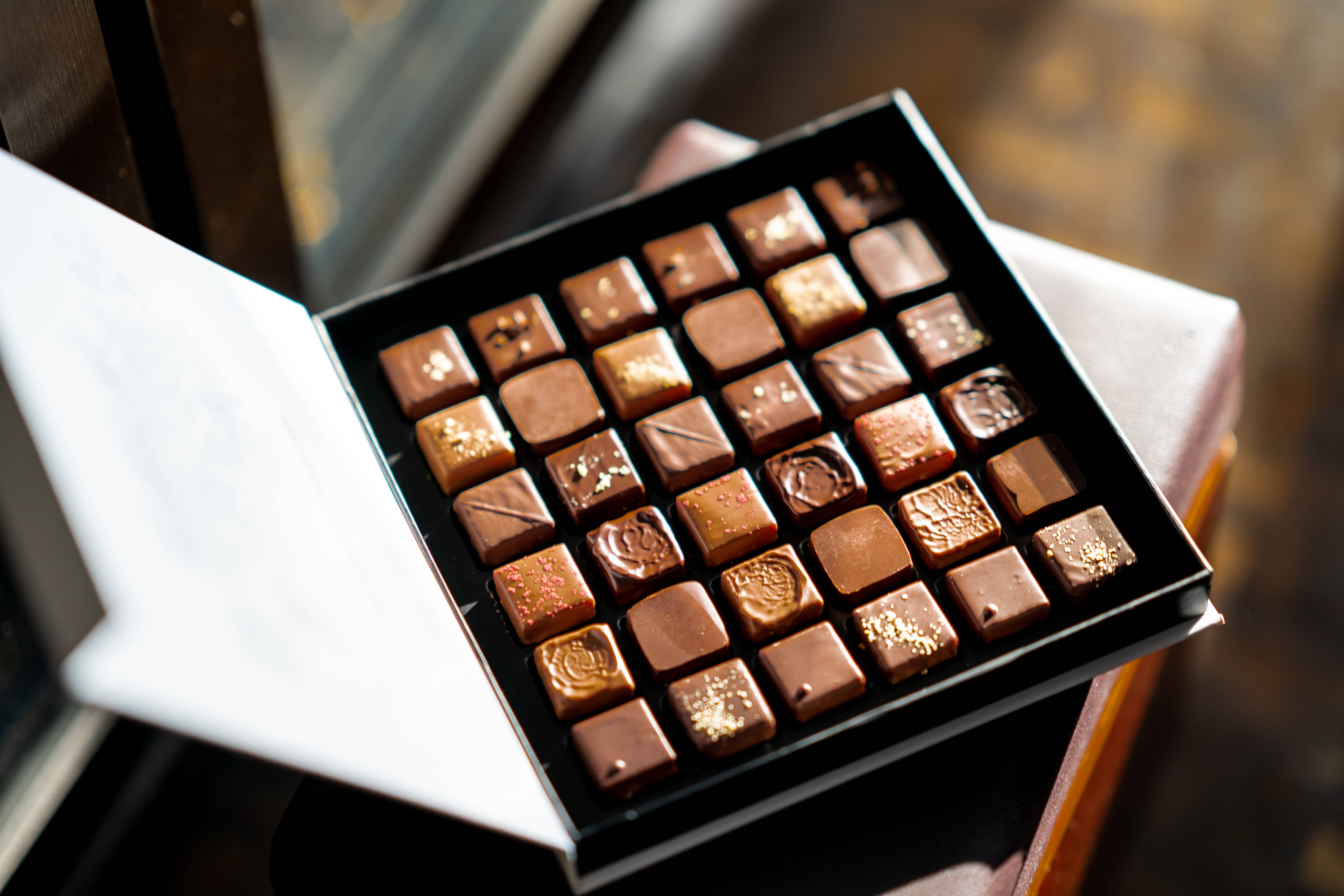 Chocolate 桌布 壁紙 — 一個豪華的打開盒子，裡面裝有各式各樣的方形巧克力，帶有金粉和紋理塗層，放在窗邊的木質表面上。