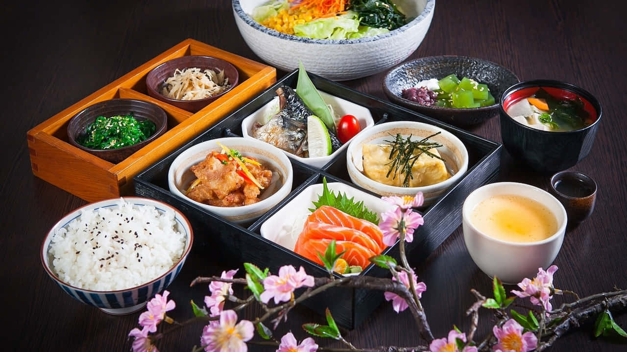 720p Food 桌布 壁紙 — 一個豪華的日本便當盒，裡面有壽司、生魚片、米飯和湯，周圍環繞著櫻花，放在深色木桌上。
