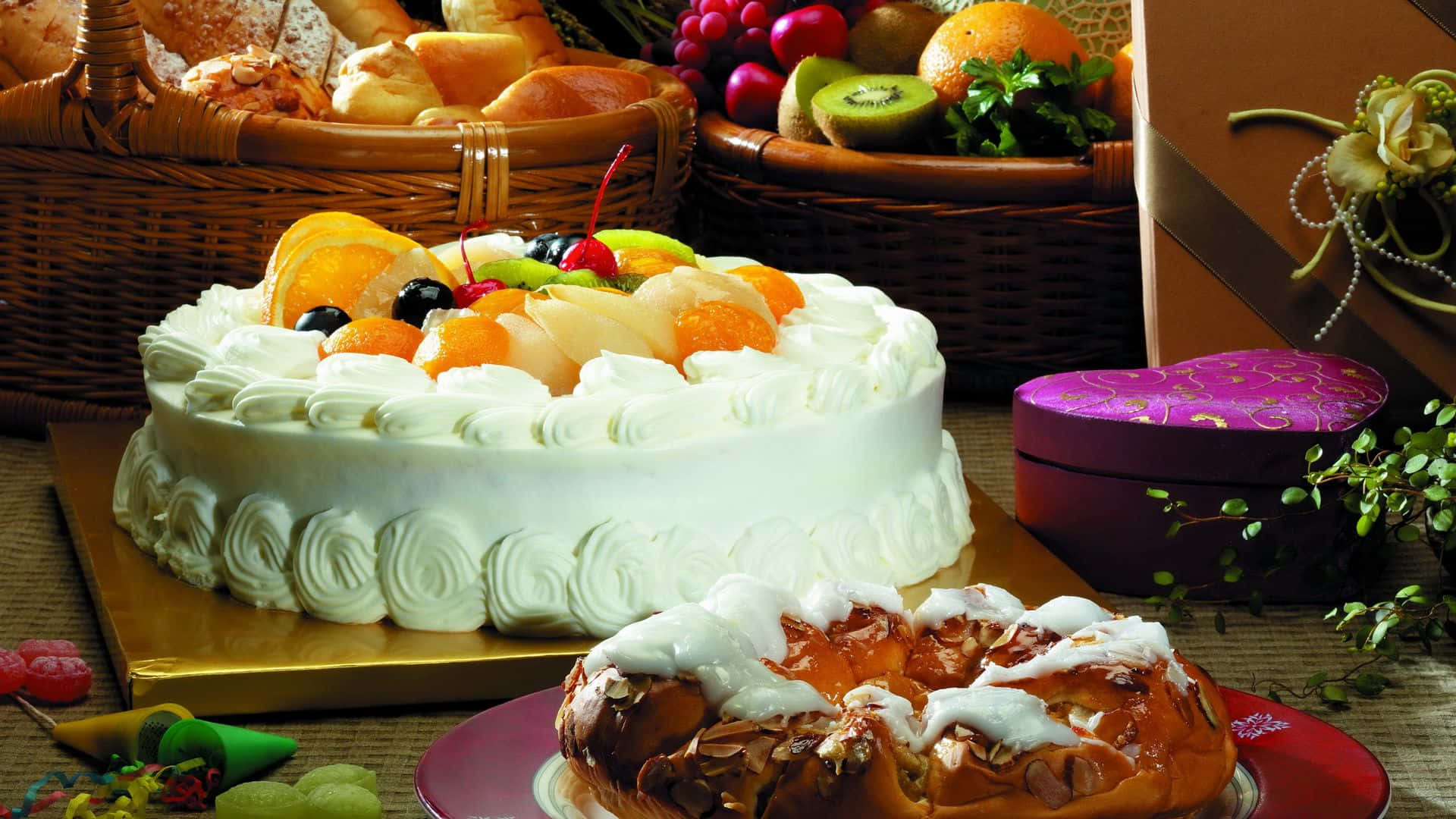Desserts 桌布 壁紙 — 一個豪華的白色糖霜蛋糕，上面放著新鮮水果，旁邊有一個辮子狀的糕點，周圍是裝滿麵包和色彩繽紛的農產品的籃子。