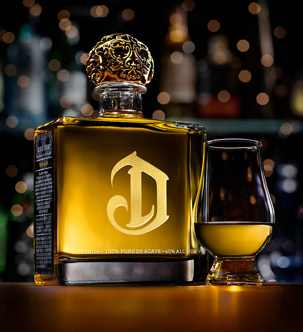 bottle 桌布 壁紙 — 一個豪華金色龍舌蘭酒瓶，帶有華麗的瓶蓋，旁邊放著一個裝滿的品酒杯，背景是條紋燈光。
