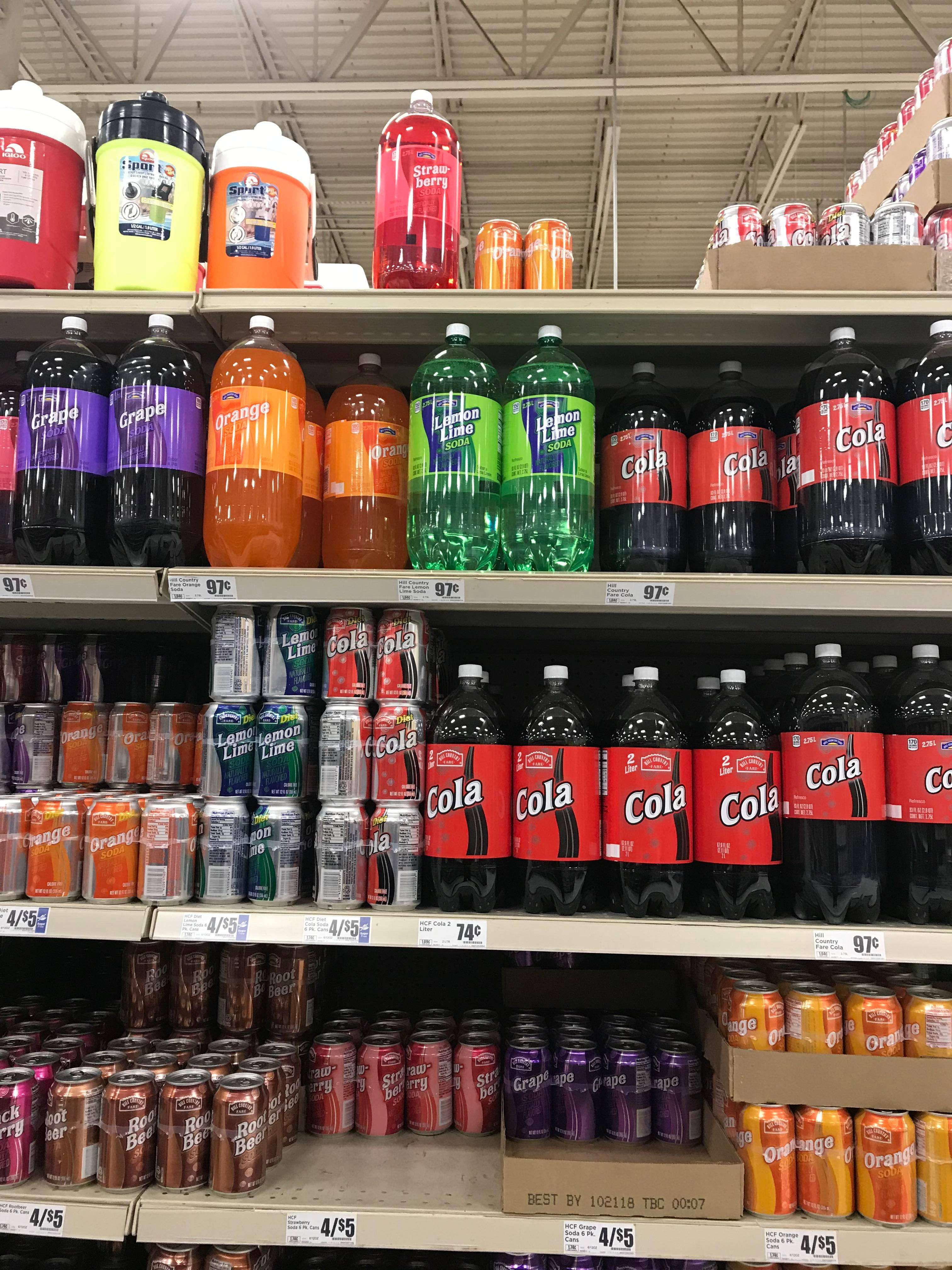 drinks 桌布 壁紙 — 一個貨品齊全的超市貨架展示著一排排色彩繽紛的汽水瓶和罐頭，在螢光燈下呈現鮮豔色調。