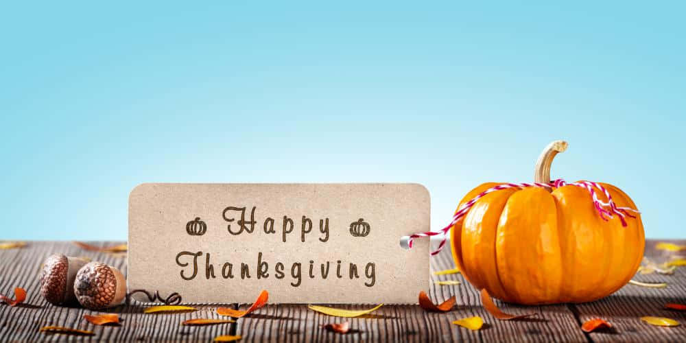 Fall Thanksgiving 桌布 壁紙 — 一個質樸的「感恩節快樂」標誌，旁邊有一個鮮豔的橙色南瓜和橡果，放在木質表面上，散落著秋天的落葉，背景是清澈的藍天。