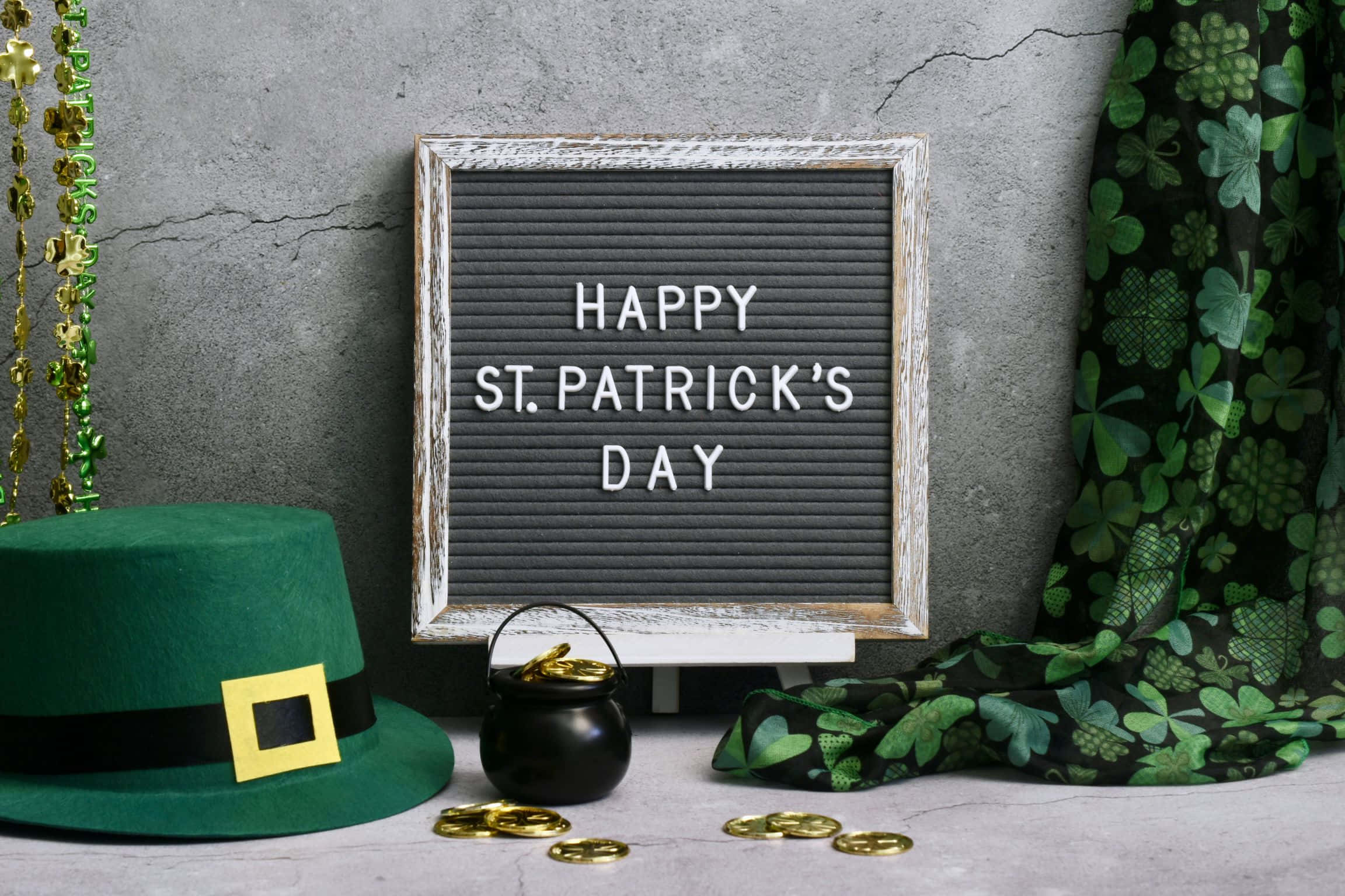 St Patrick's Day 桌布 壁紙 — 一個質樸的字板顯示「HAPPY ST. PATRICK'S DAY」，周圍環繞著綠色帽子、金幣和三葉草布料，背景是裂開的混凝土牆。