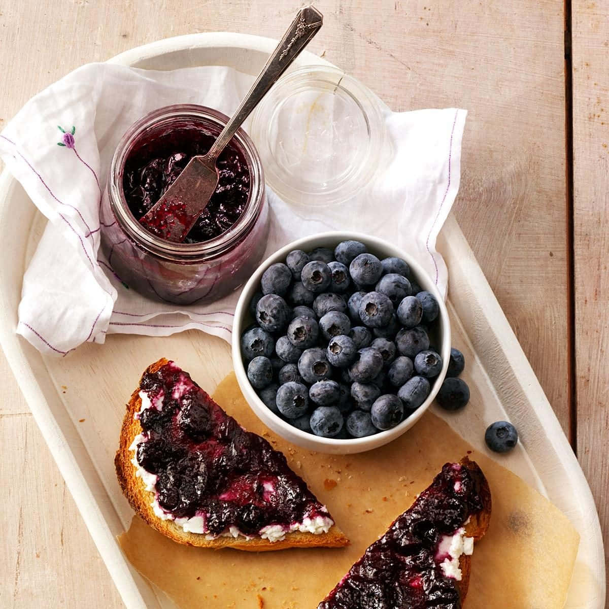 Blueberry Jam 桌布 壁紙 — 一個質樸的托盤上放著新鮮藍莓、一瓶自製藍莓果醬和塗上奶油及果醬的吐司片，置於木桌上。