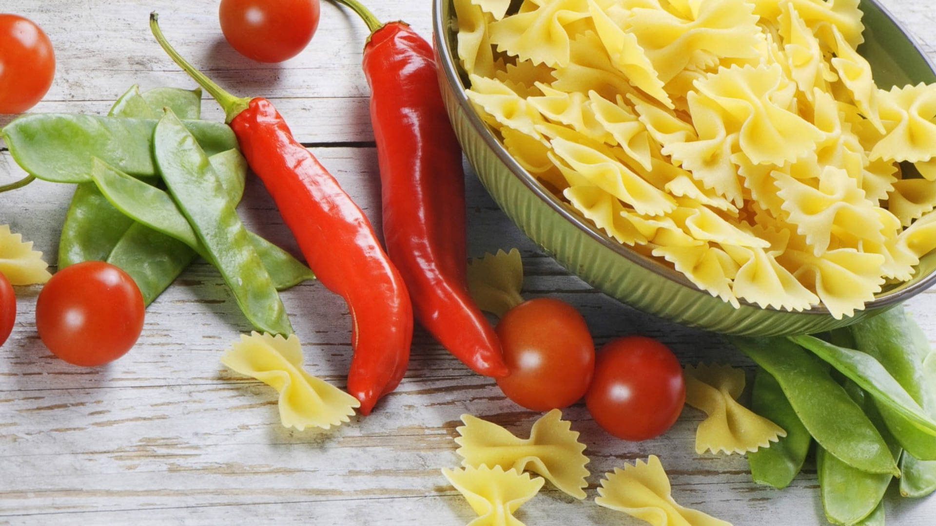 Pasta 桌布 壁紙 — 一個質樸的木桌上展示一碗蝴蝶結義大利麵，周圍是新鮮的紅辣椒、櫻桃番茄和綠色荷蘭豆。