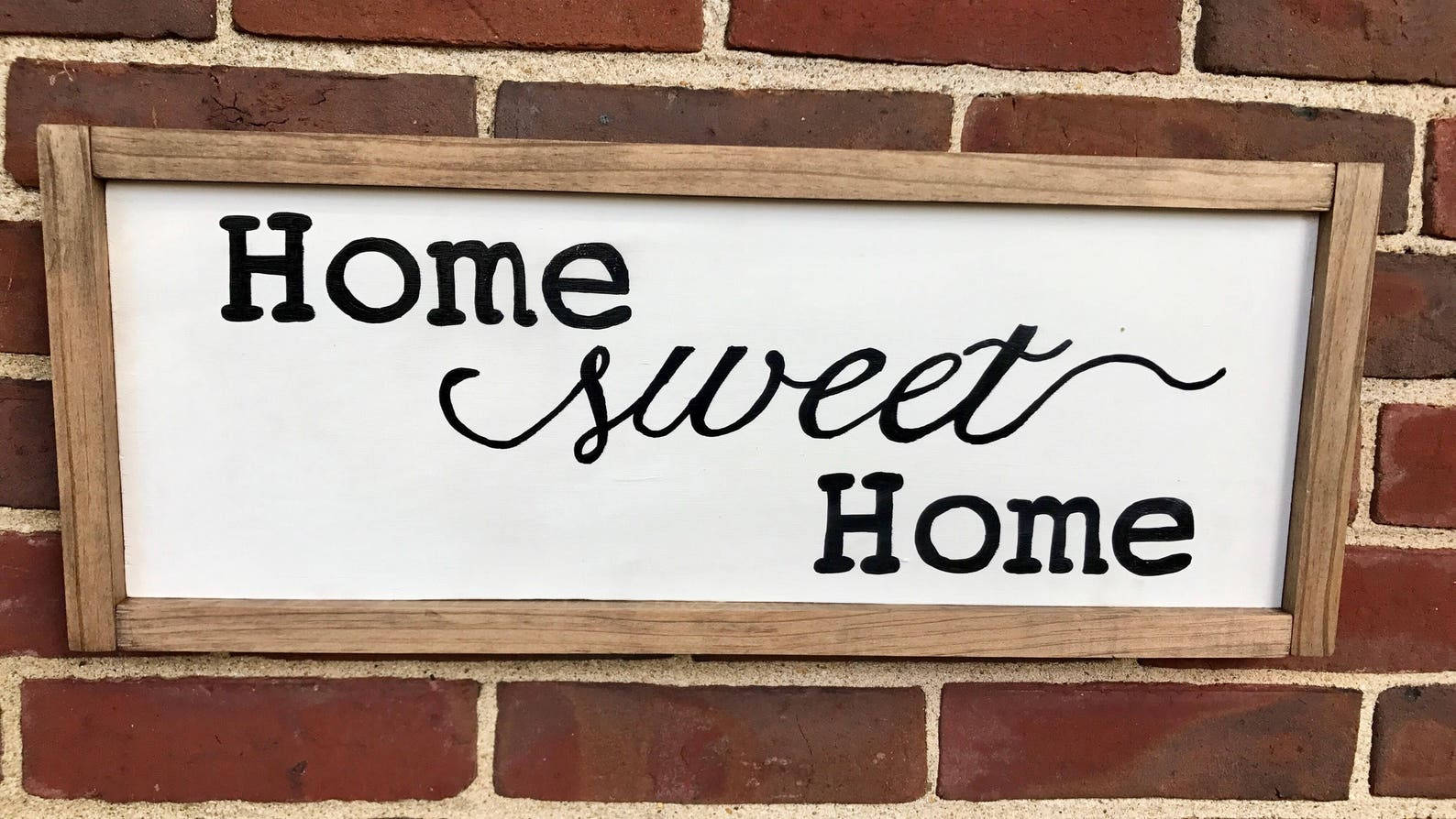 甜蜜的家 桌布 壁紙 — 一個質樸的木牌，上面用黑色草書寫著「Home sweet Home」，掛在紅磚牆上。