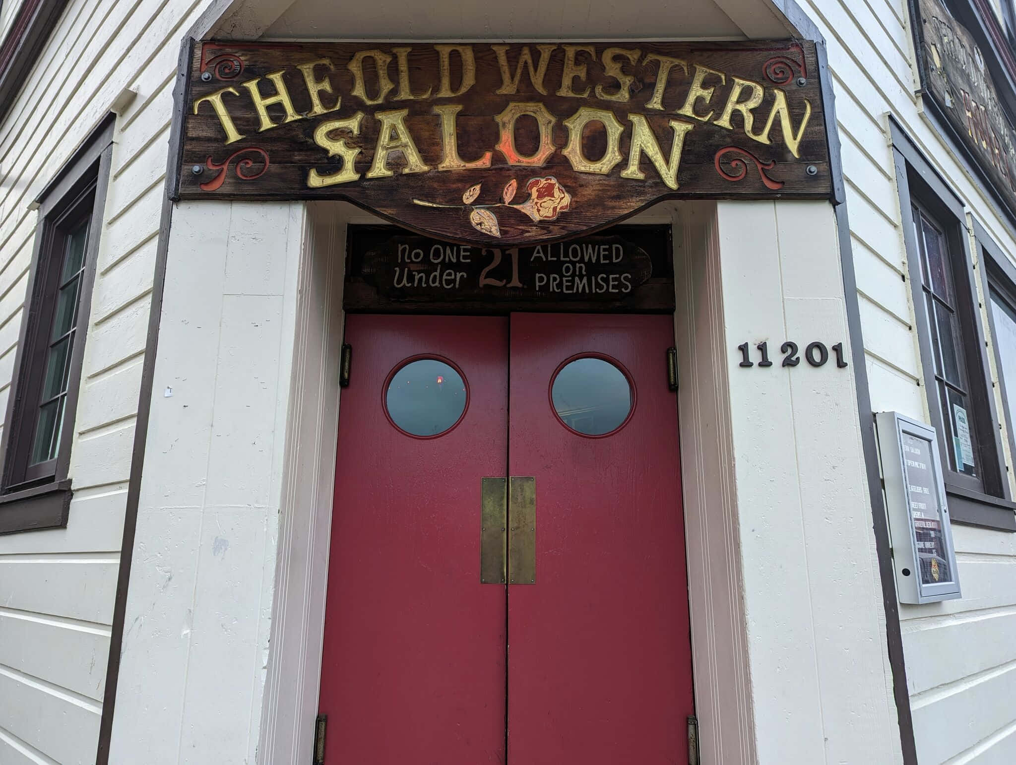 Old Western 桌布 壁紙 — 一個質樸的木牌寫著「The Old Western Saloon」，位於白色木板建築上帶有圓窗的紅色雙門上方。