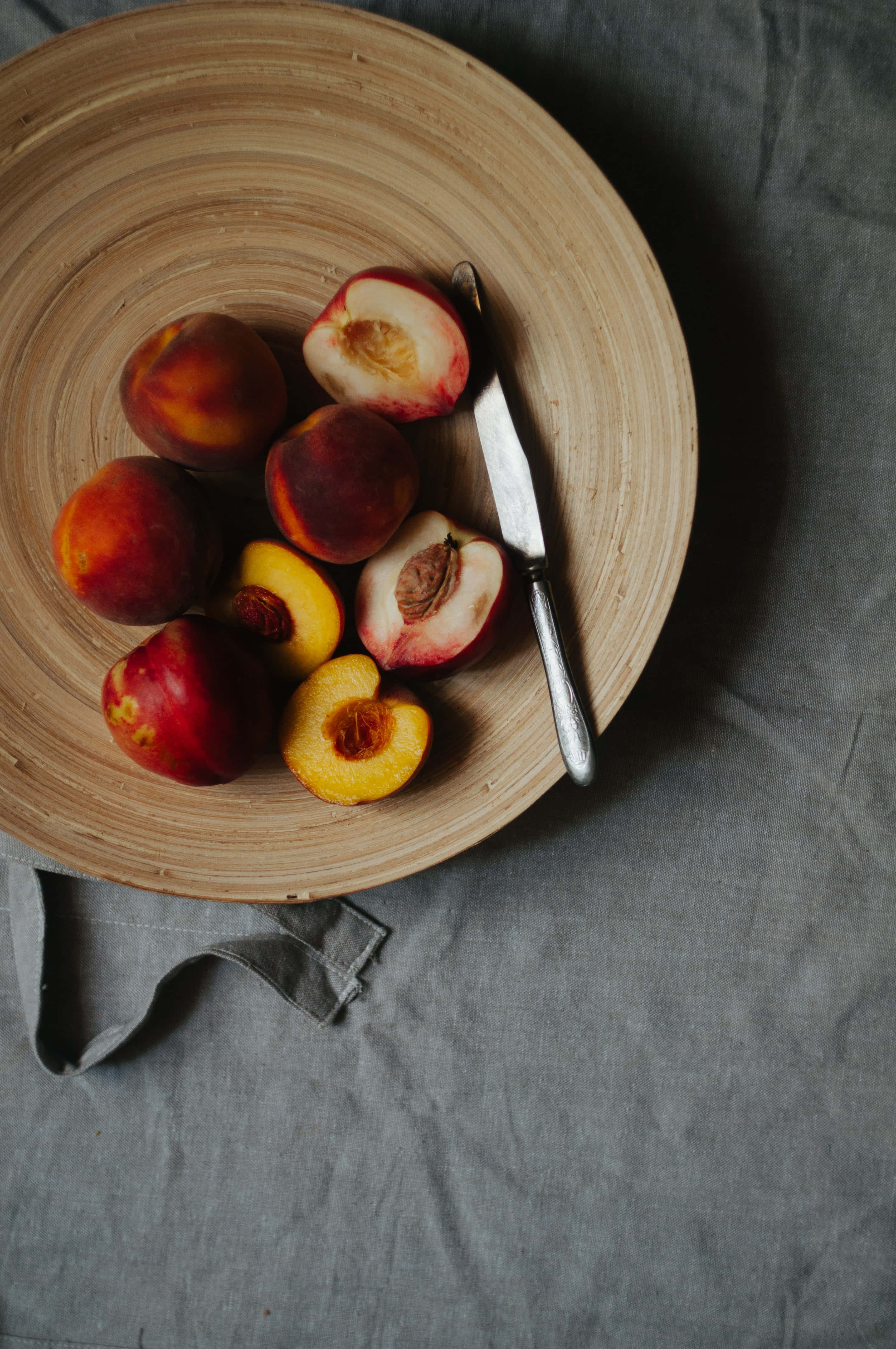 Peaches 桌布 壁紙 — 一個質樸的木盤上放著成熟的桃子和一把刀，置於灰色亞麻布上，柔和自然光下。