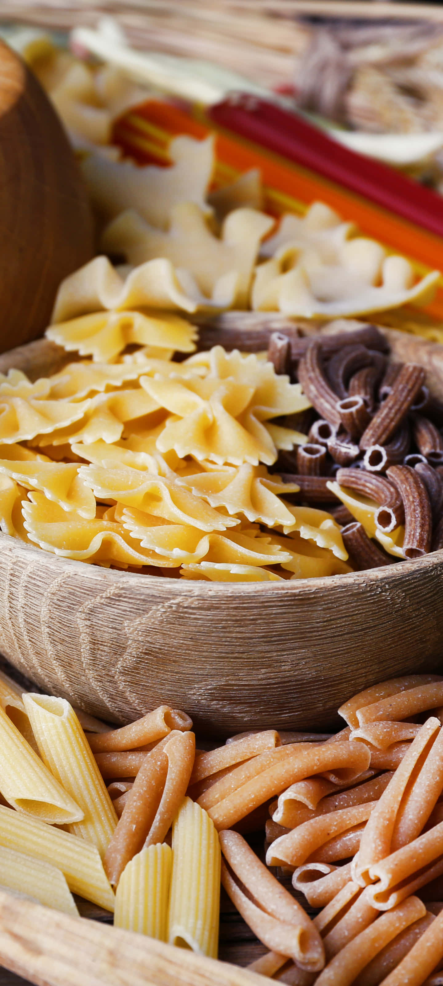 Best Pasta 桌布 壁紙 — 一個質樸的木碗，裝滿了色彩繽紛的義大利麵種類，包括蝴蝶麵和筆管麵，呈現溫暖的大地色調。