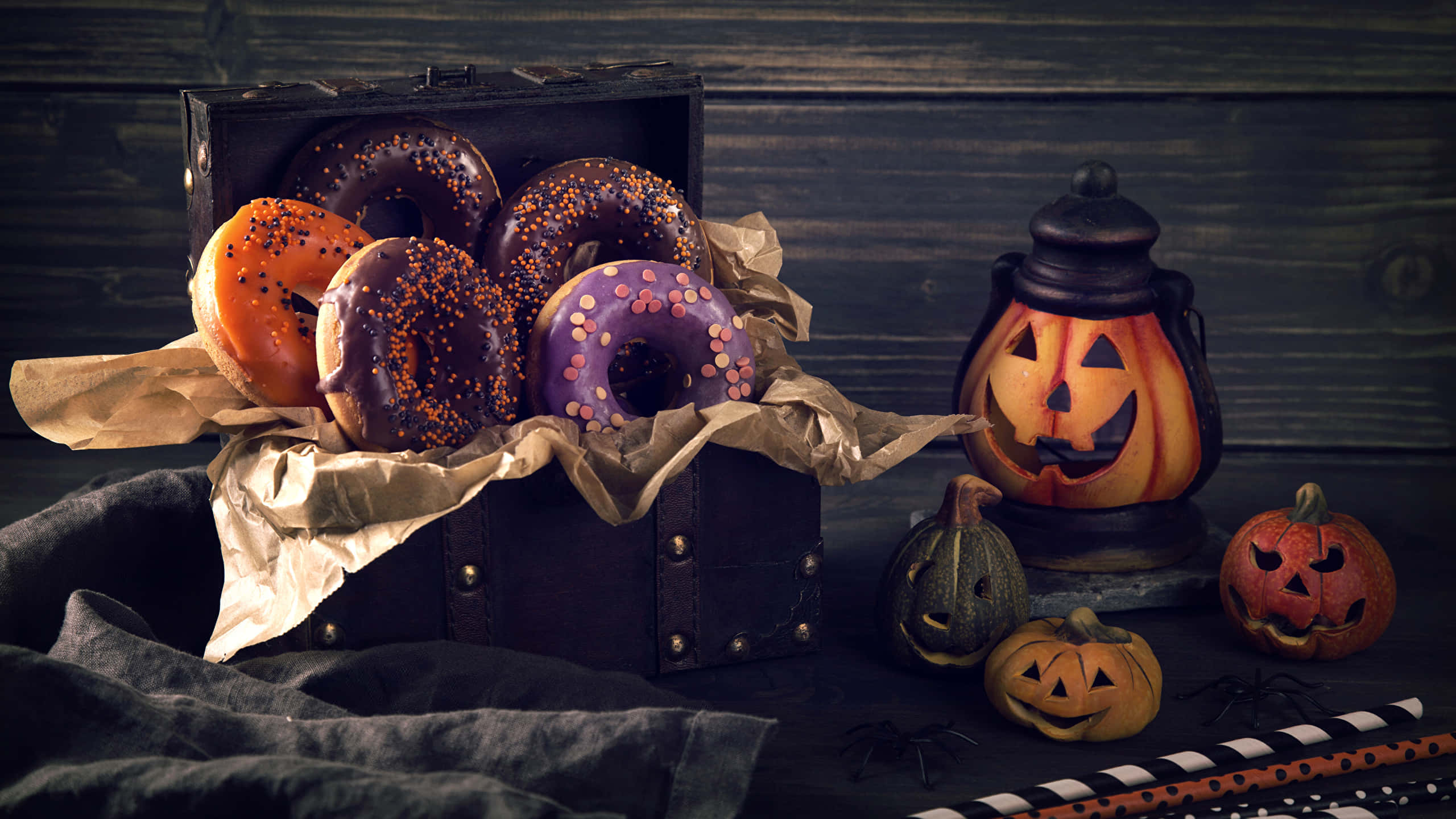 Halloween Treats 桌布 壁紙 — 一個質樸的木箱裝滿色彩繽紛的萬聖節甜甜圈，旁邊有一個發光的傑克南瓜燈和雕刻南瓜，放在深色木質表面上。
