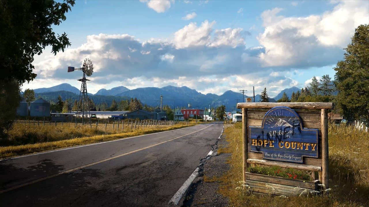 Far Cry 5 桌布 壁紙 — 一個質樸的木製Hope County標誌矗立在鄉村道路上，背景有山脈和風車，天空部分多雲。