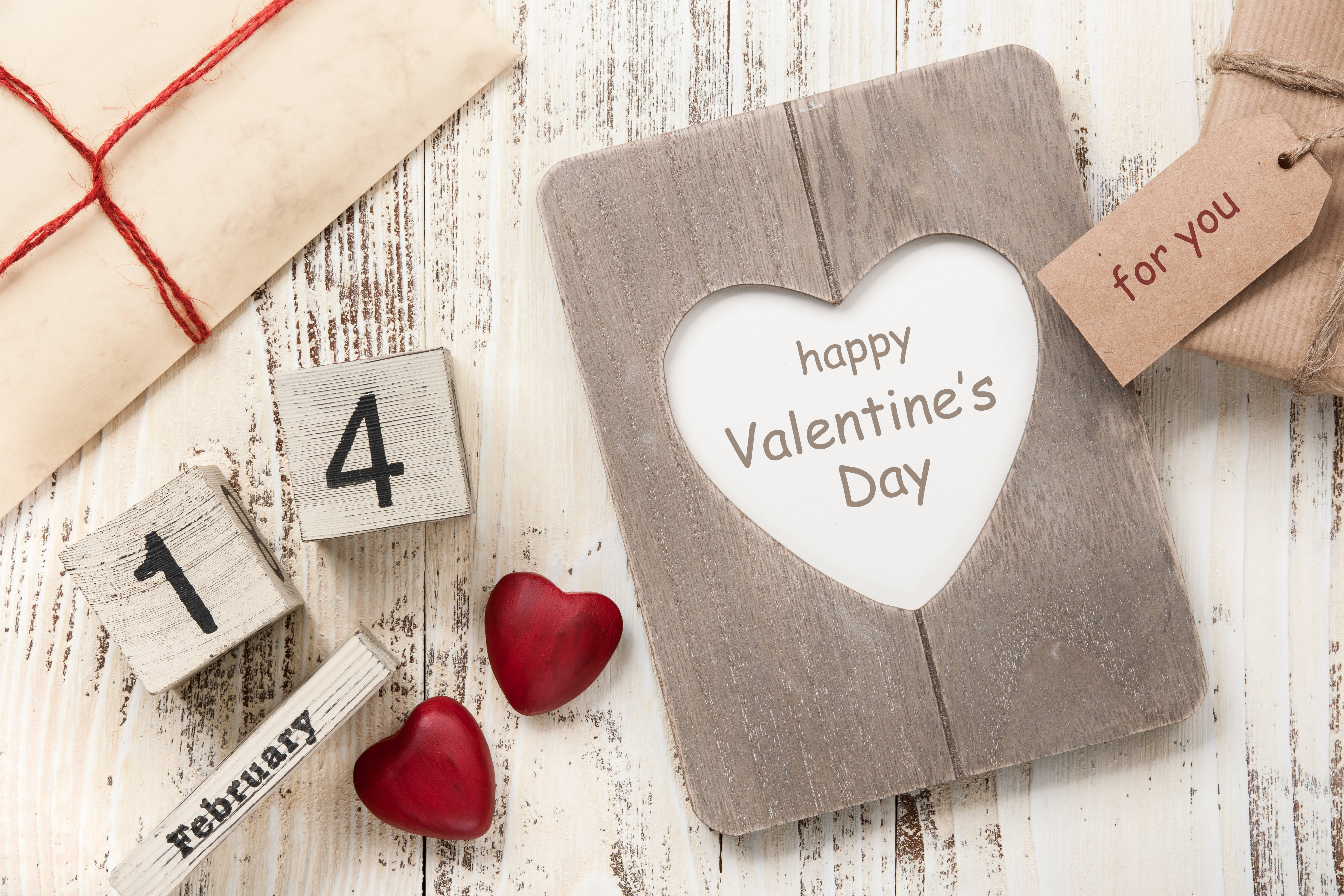 情人節 桌布 壁紙 — 一個質樸的木製心形相框，上面有「happy Valentine's Day」文字，旁邊有紅色心形、日期方塊和禮物，放在經風化的白色木質表面上。