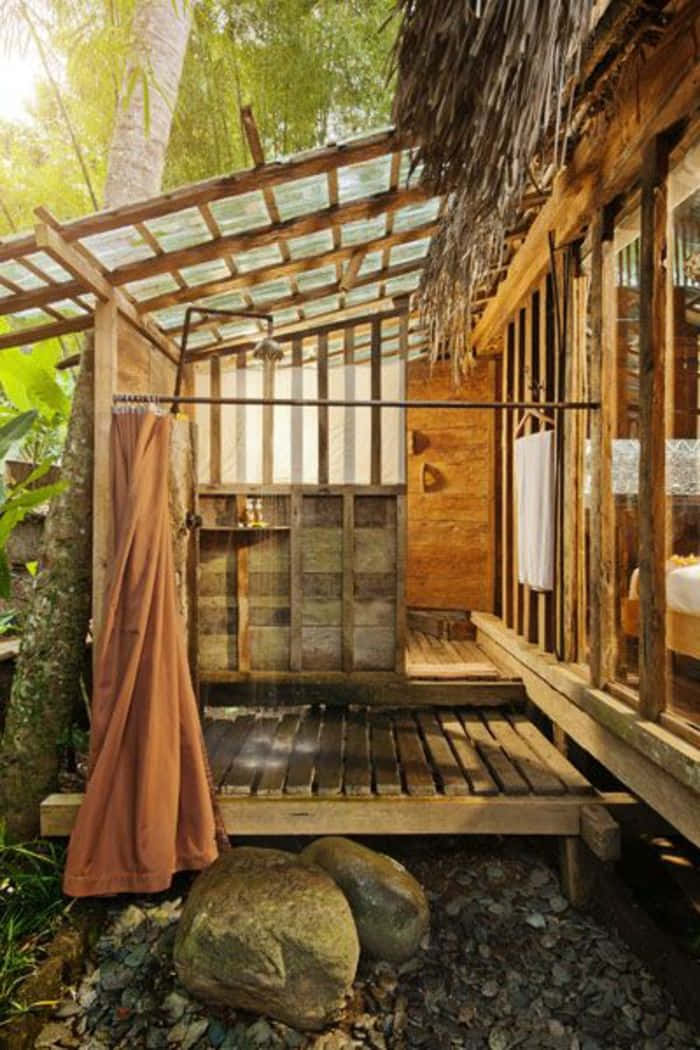 cabin 桌布 壁紙 — 一個質樸的木製戶外淋浴間，隱藏在茂密的熱帶植物中，配有茅草屋頂和石頭地面。