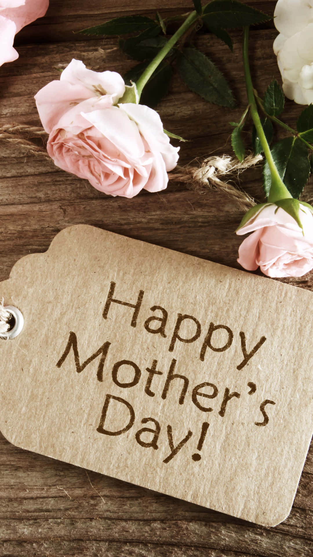 Mother's Day 桌布 壁紙 — 一個質樸的木製標籤，上面寫著 Happy Mother's Day，周圍環繞著柔和的粉紅色和白色玫瑰，放在經風化的木質表面上。