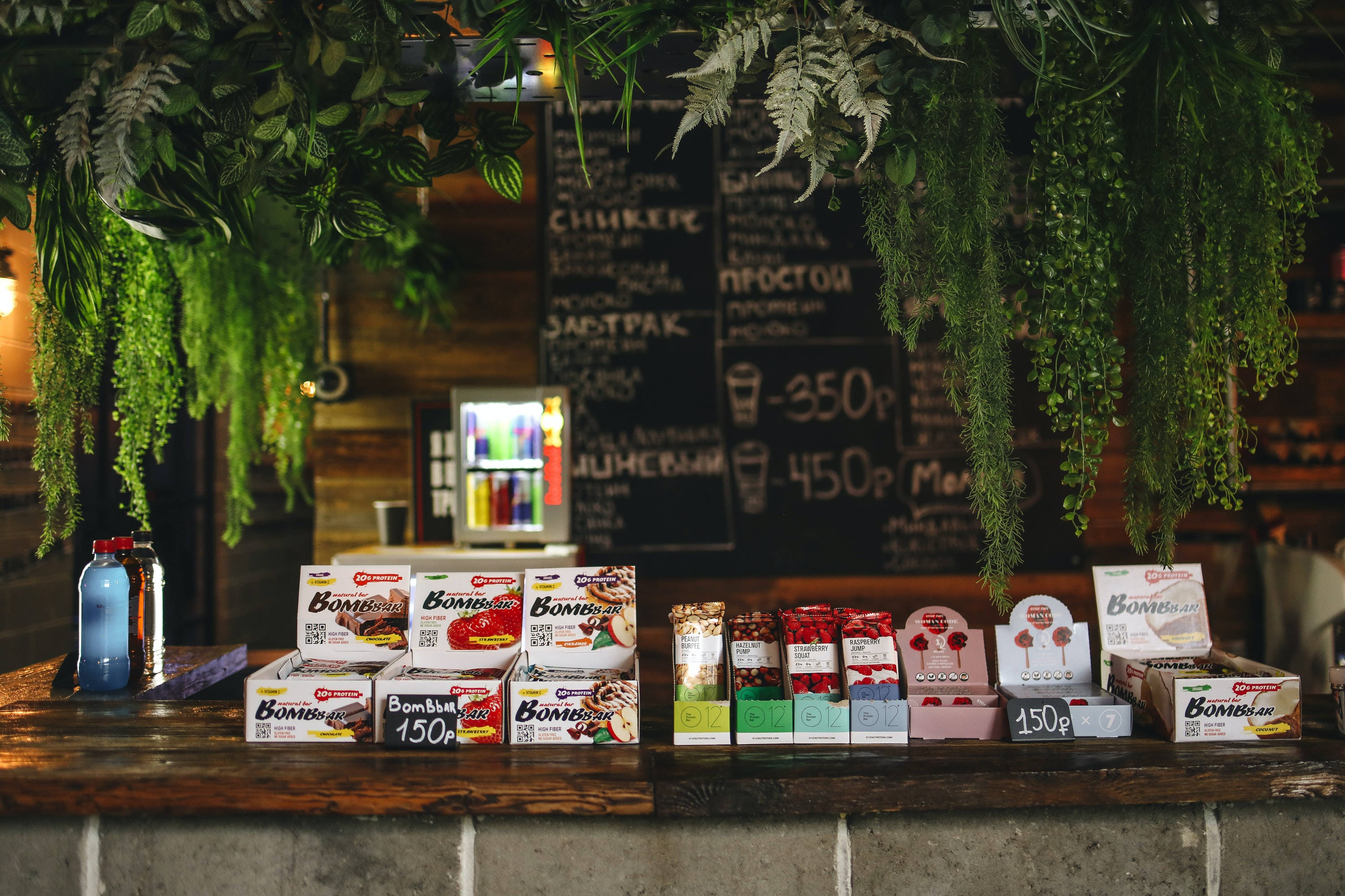 Coffee Shop 桌布 壁紙 — 一個質樸的木製櫃檯展示著色彩繽紛的零食盒，上方懸掛著茂盛的蕨類植物，背景是黑板菜單。