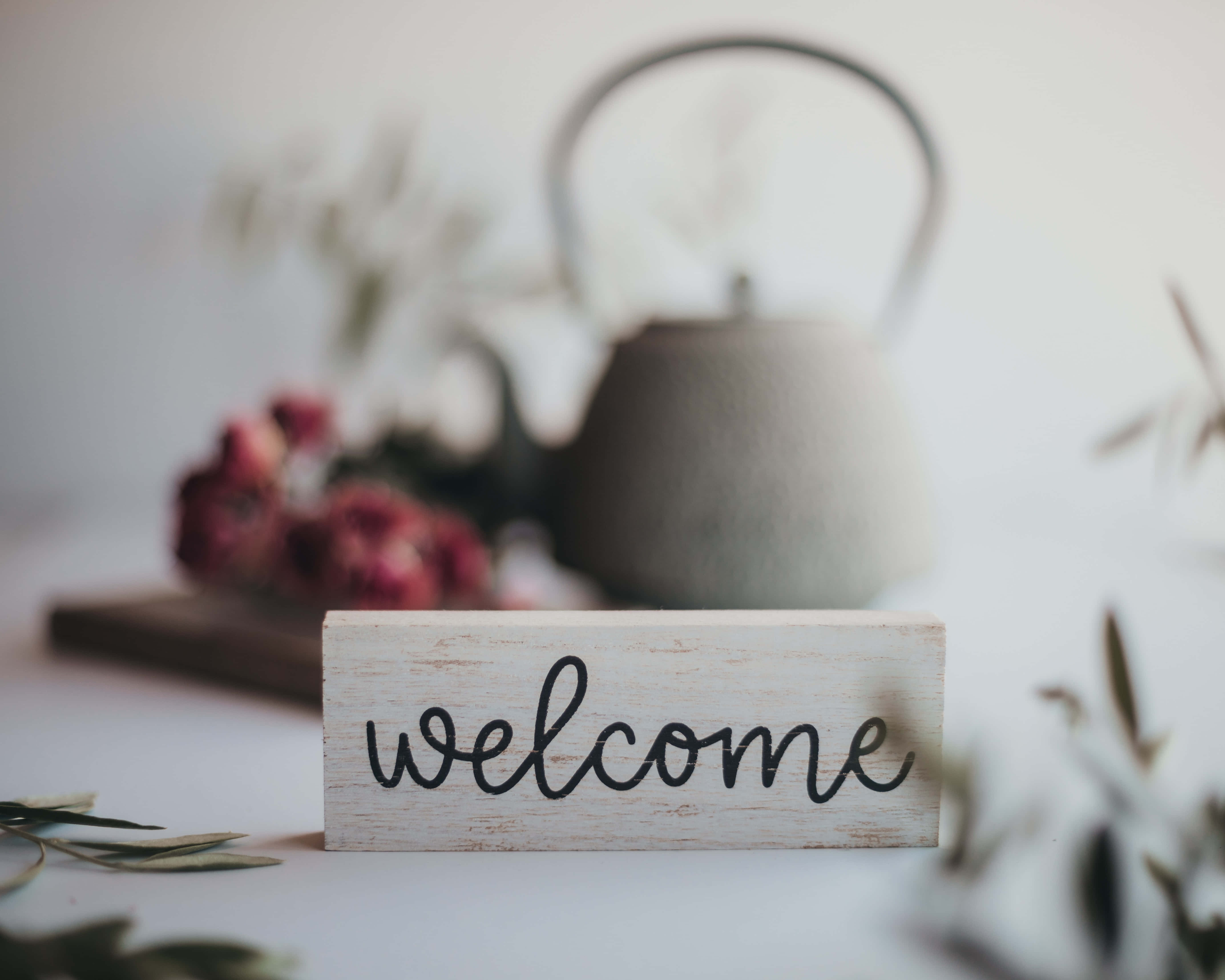 Welcome 桌布 壁紙 — 一個質樸的木製歡迎牌放在模糊的灰色茶壺和乾花前面，背景是柔軟的白色表面。