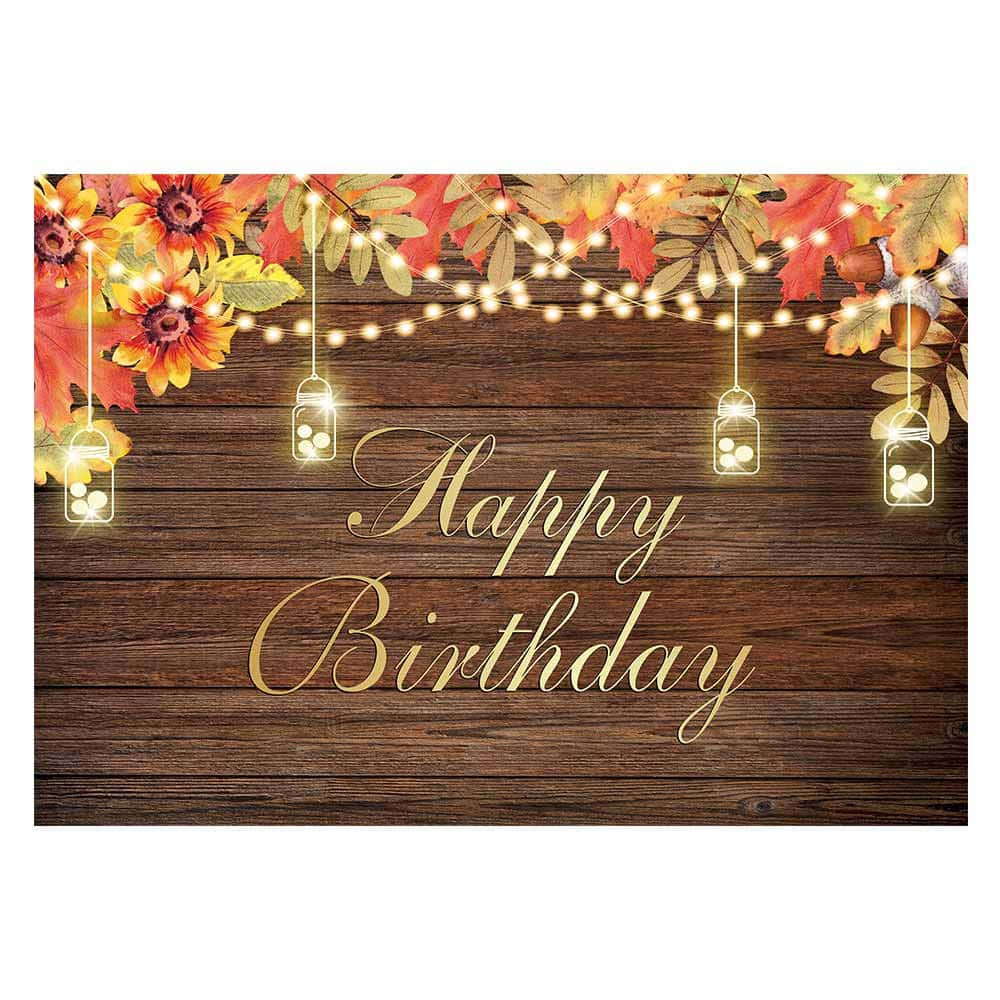 Fall Birthday 桌布 壁紙 — 一個質樸的木製背景，裝飾著秋葉、向日葵和發光的串燈，配上梅森罐燈籠，展示金色草書文字Happy Birthday。